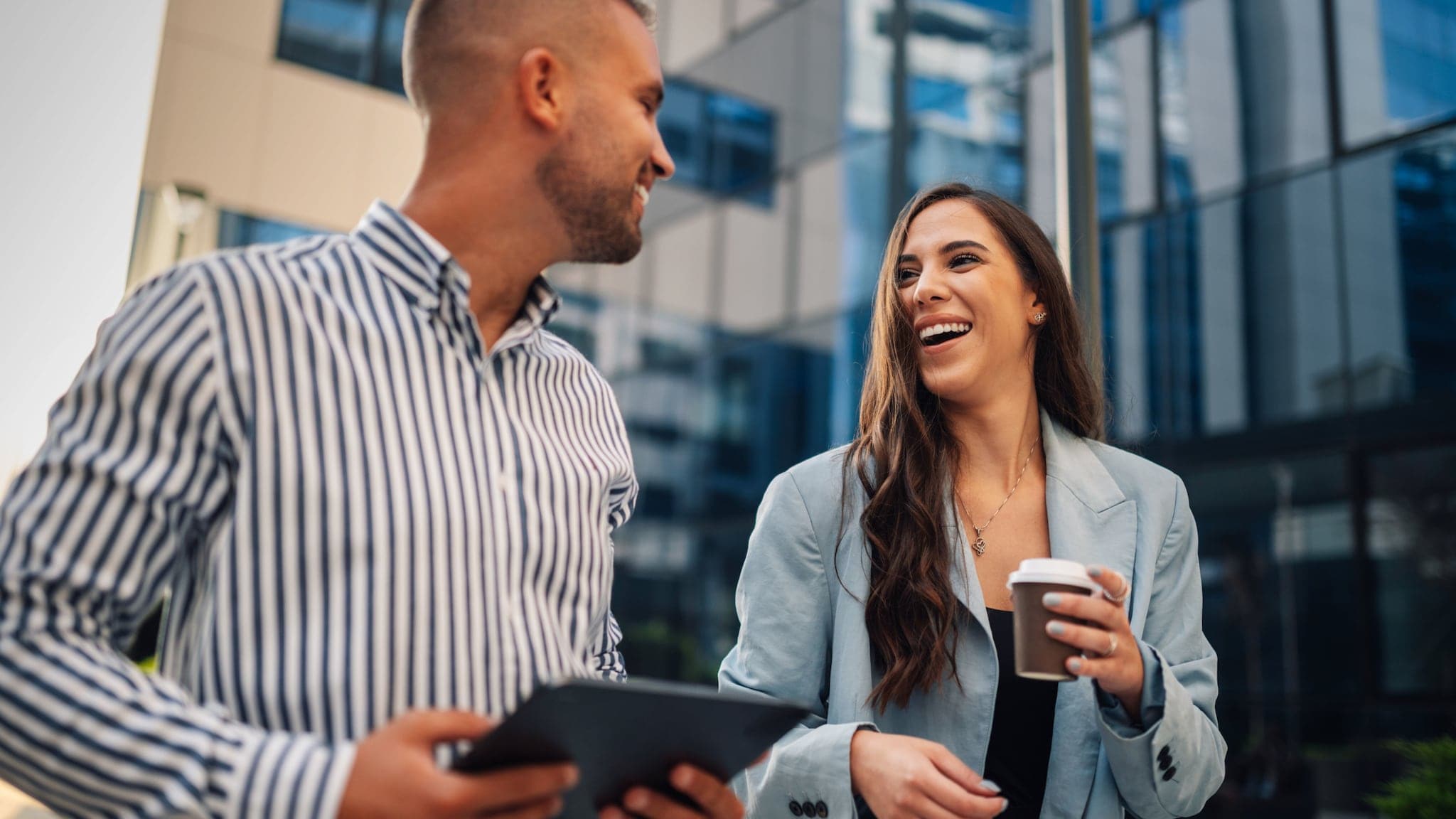 Eine Frau mit Kaffeebecher und ein Mann mit Tablet in den Händen unterhalten sich gut gelaunt vor einem Bürogebäude. Eine Frau mit Kaffeebecher und ein Mann mit Tablet in den Händen unterhalten sich gut gelaunt vor einem Bürogebäude.