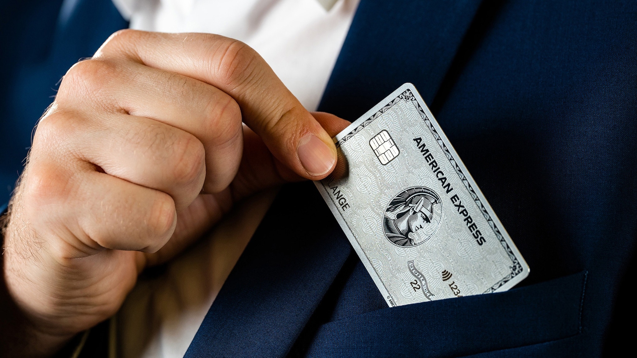 Nahaufnahme einer männlichen Hand, die eine silberne Kreditkarte von American Express aus der Brusttasche eines blauen Jacketts zieht.