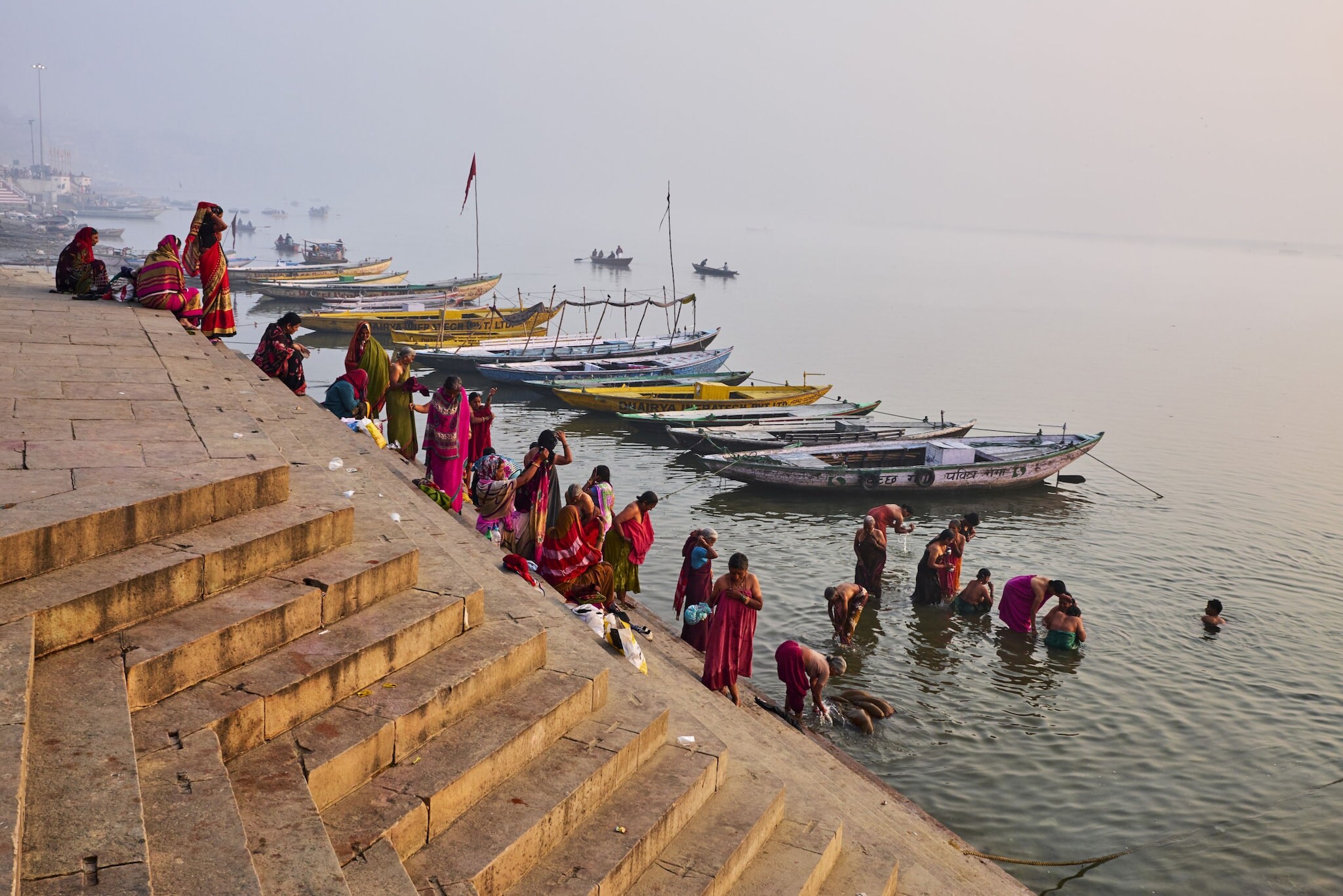 Stufenartige Uferbefestigung am Ganges mit in roten Umhängen gekleideten Menschen und Boote im Wasser.
