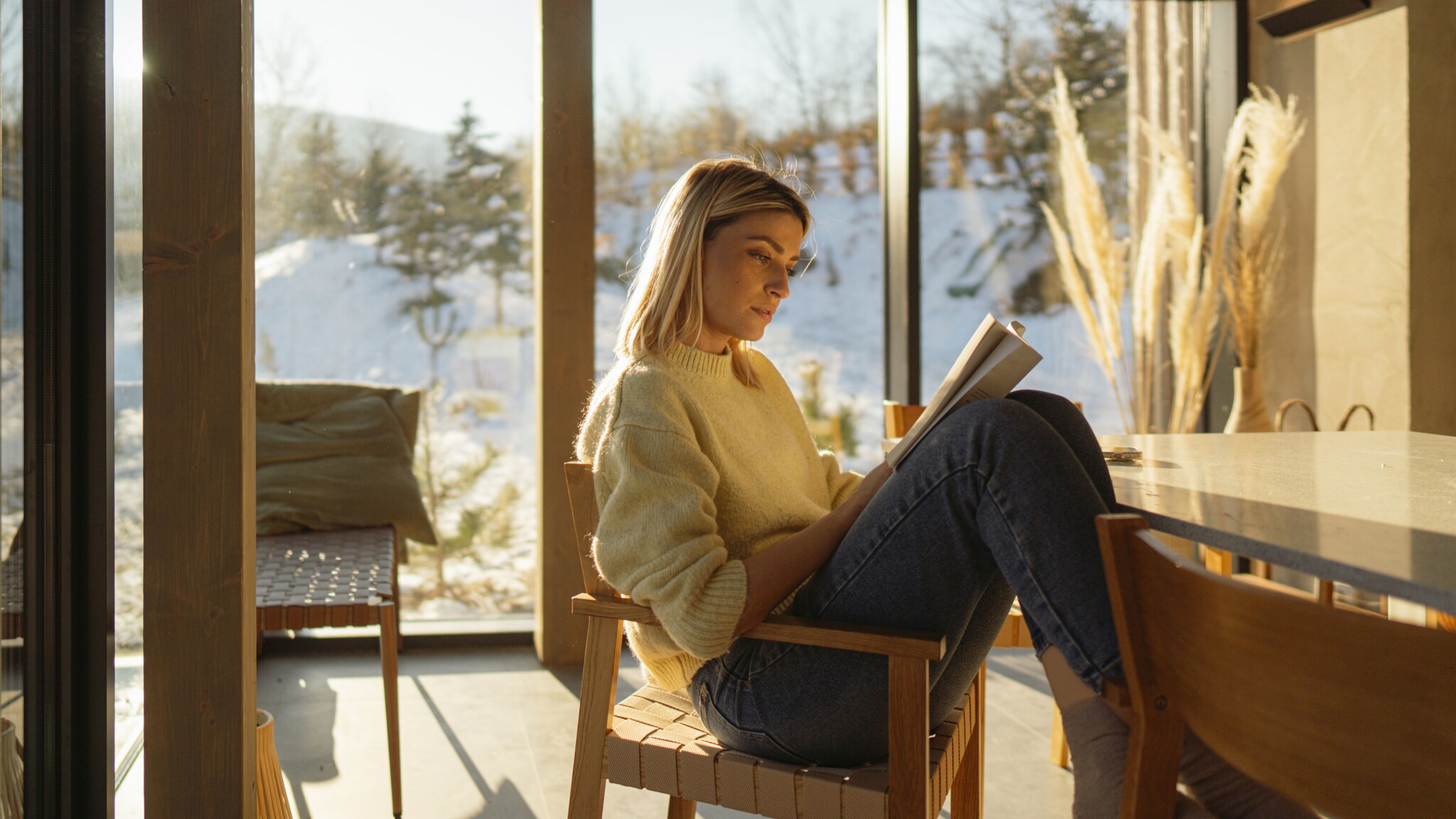 Eine Frau liest mit angewinkelten Beinen auf einem Stuhl in einem Raum mit bodentiefen Fenstern vor einer verschneiten Landschaft.