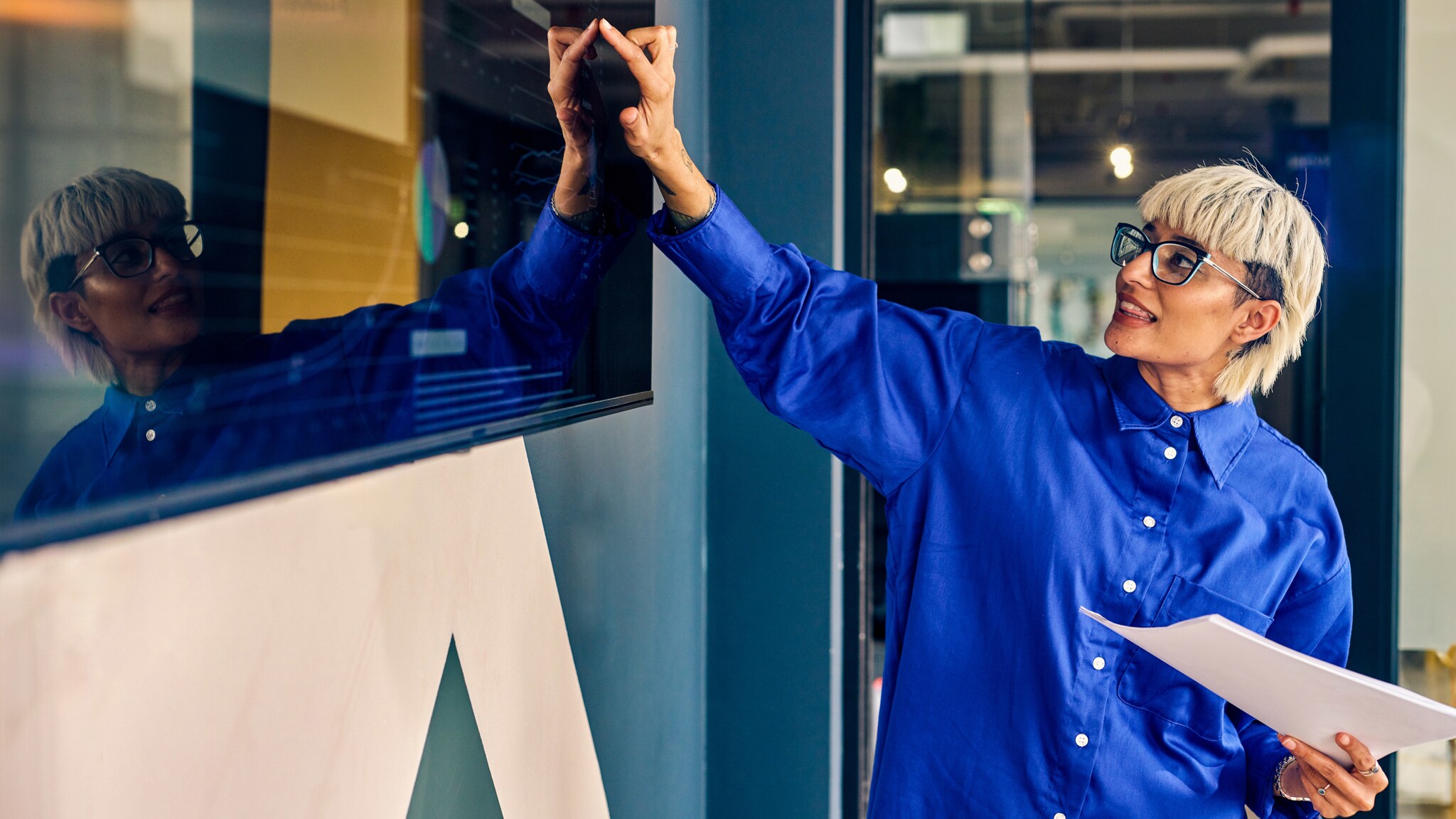 Eine Frau mit Wolf-Cut im blauen Hemd zeigt auf ein großes Display, in der anderen Hand hält sie Papiere. Eine Frau mit Wolf-Cut im blauen Hemd zeigt auf ein großes Display, in der anderen Hand hält sie Papiere.