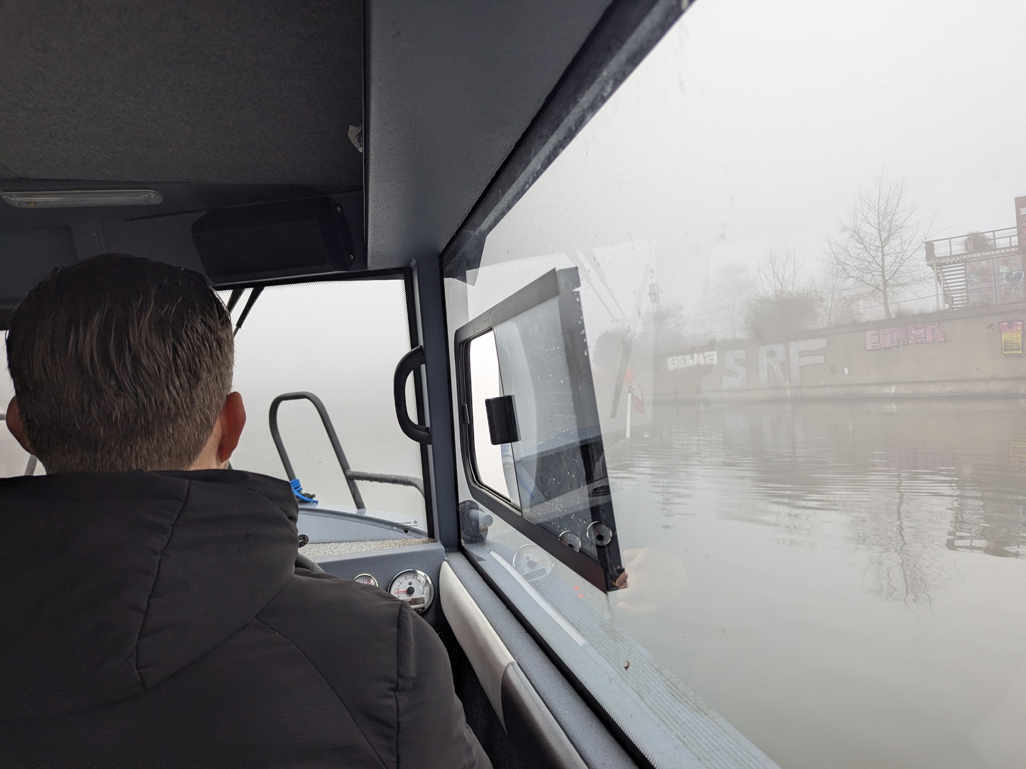 Blick von hinten auf eine Person im Steuerhaus eines Motorboots, draußen dichter Nebel auf einer Wasserstraße.