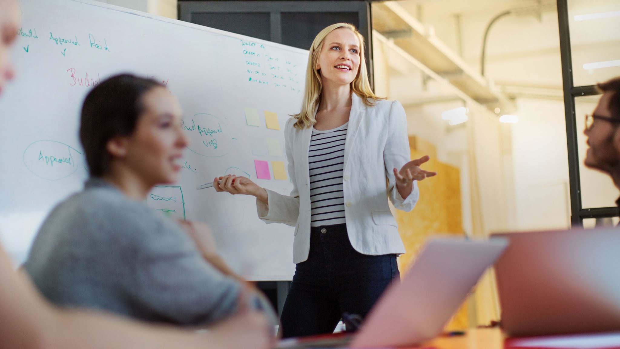 Eine Frau im weißen Blazer steht vor einem Whiteboard während einer Präsentation mit sitzenden Personen in einem Büro.