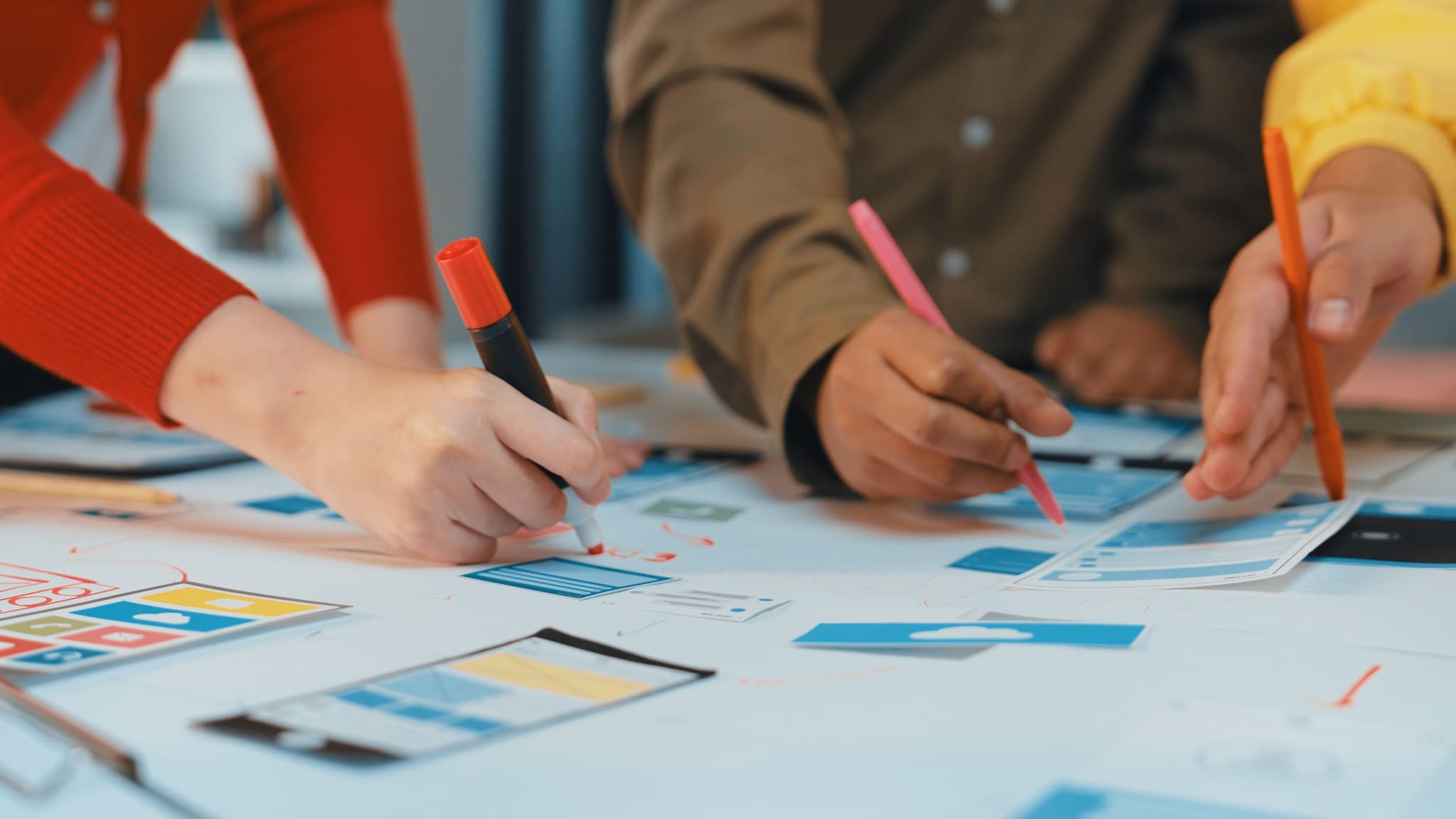 Mehrere Hände bearbeiten mit farbigen Stiften ausgedruckte Designentwürfe auf einem Tisch.