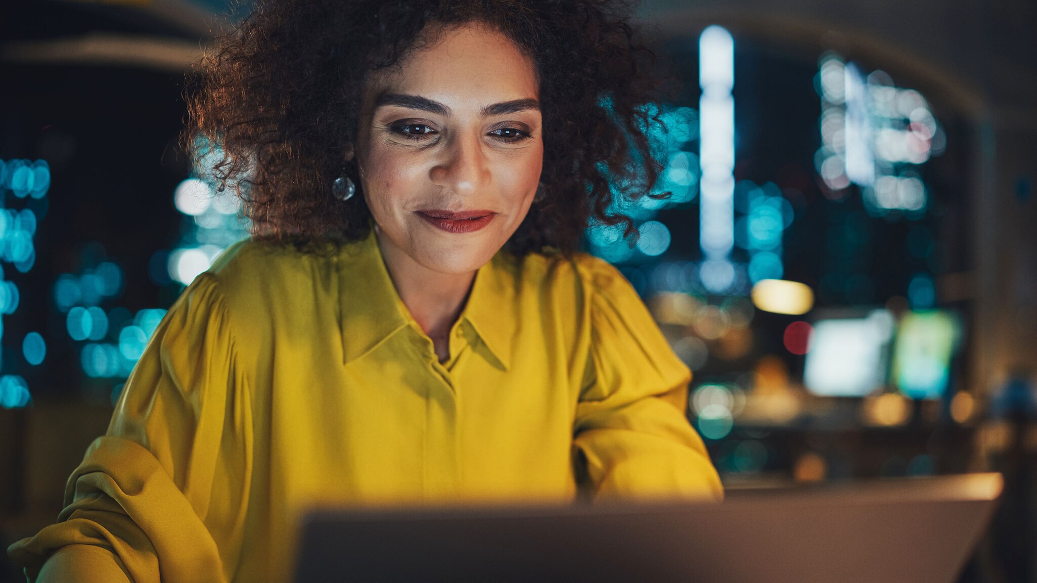 Eine Frau in gelber Bluse sitzt an einem Laptop, ihr Gesicht vom Licht des Displays beleuchtet, in einem Büro vor einer Skyline bei Nacht.