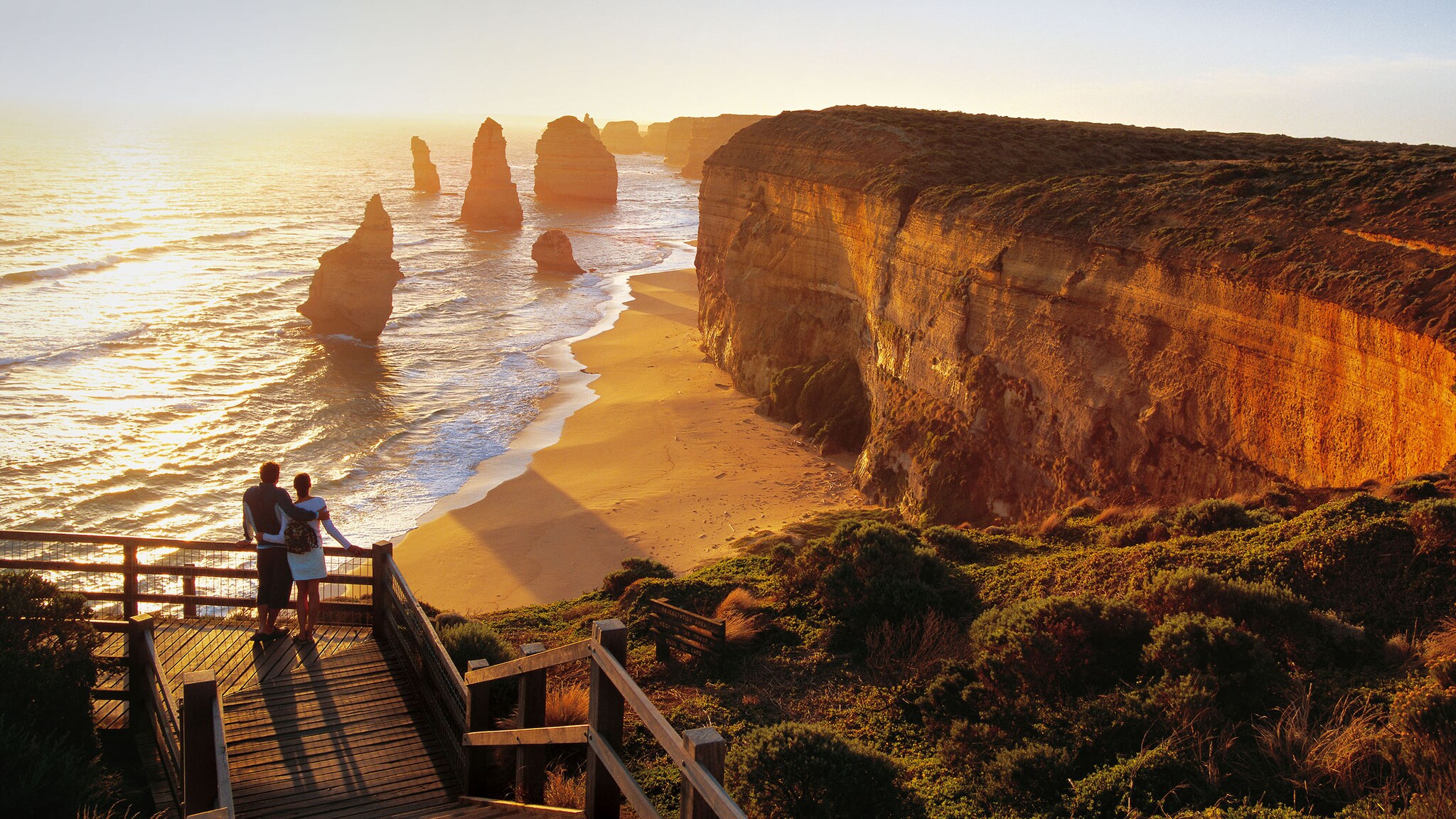 Zwei Personen stehen auf einer Aussichtsplattform aus Holz an einer Klippe am Meer im warmen Sonnenlicht.