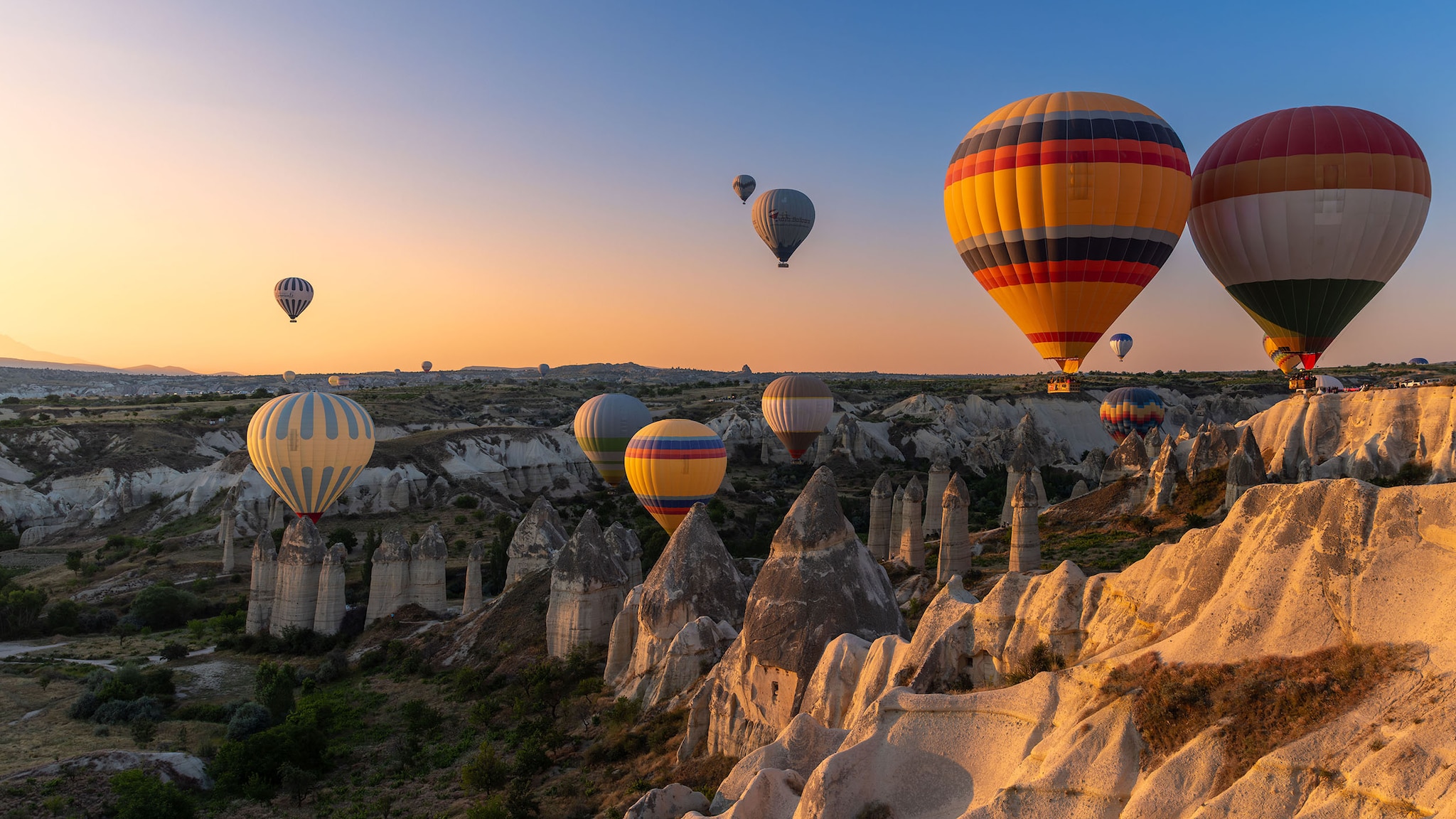 Mehrere Heißluftballons schweben über eine Tuffsteinlandschaft mit spitzen Felsformationen bei Sonnenaufgang.
