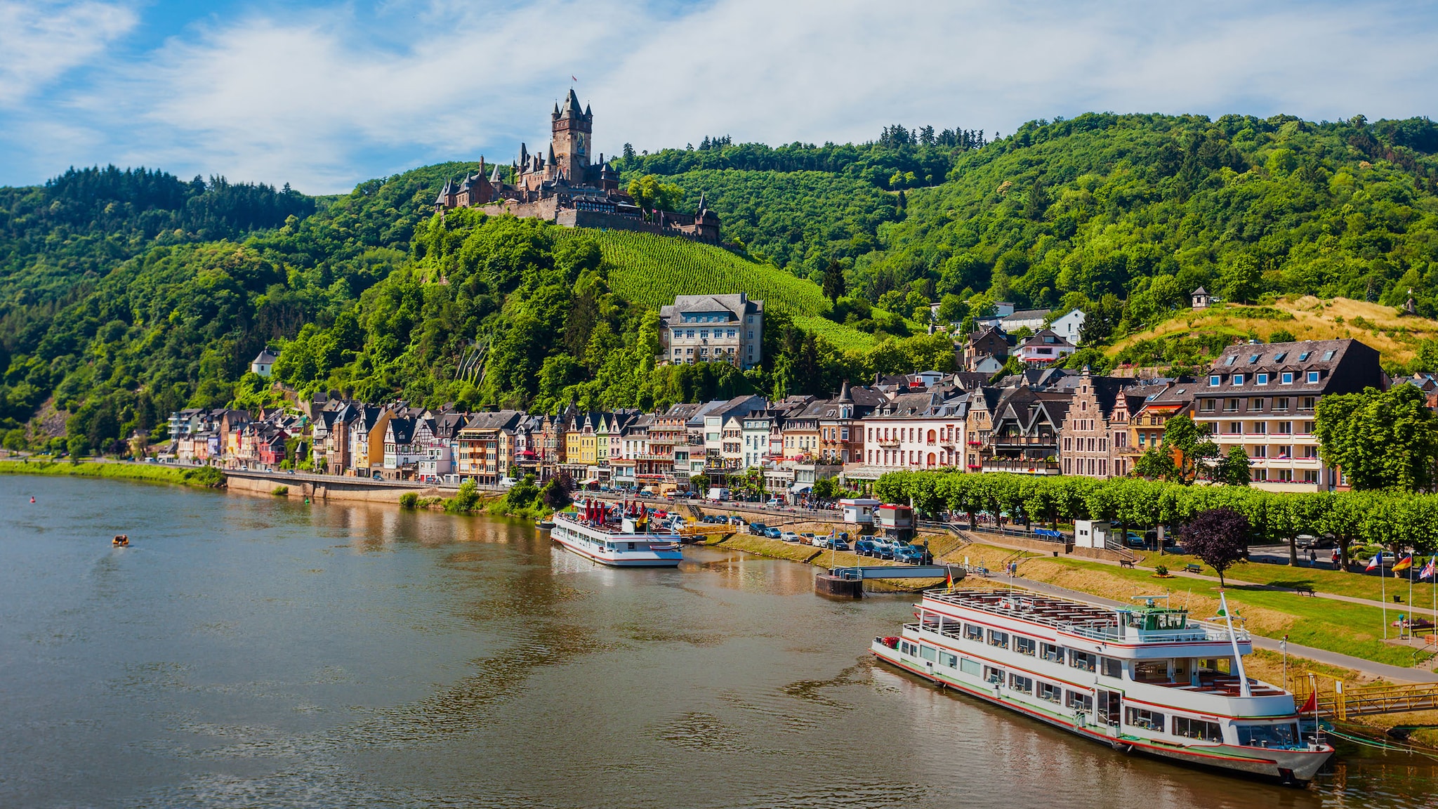 Mittelalterliche Kleinstadt am Fluss mit Ausflugsbooten vor grüner Hügellandschaft mit Burganlage unter blauem Himmel.