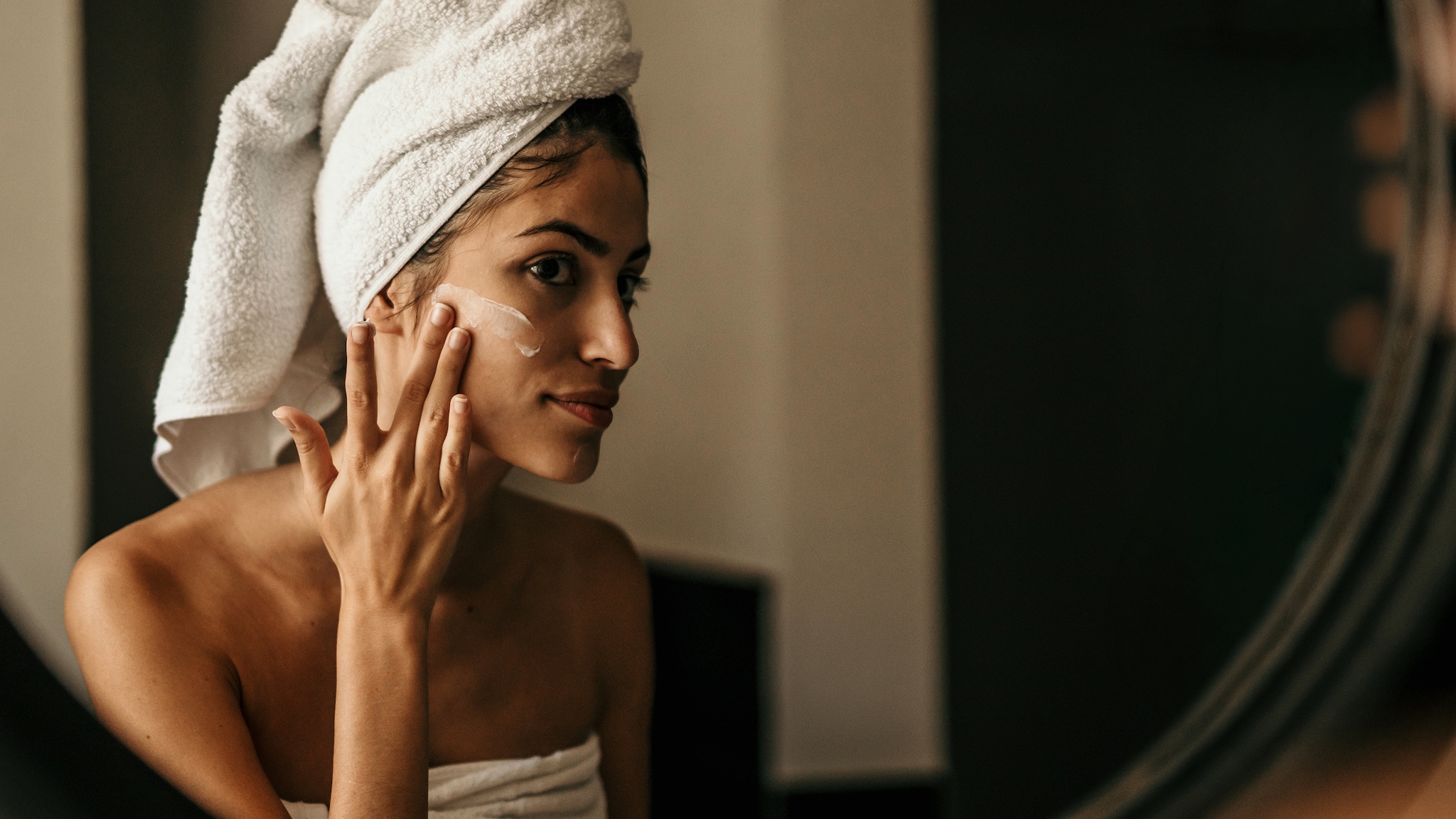 Eine Frau steht vor einem Spiegel und cremt ihr Gesicht ein.