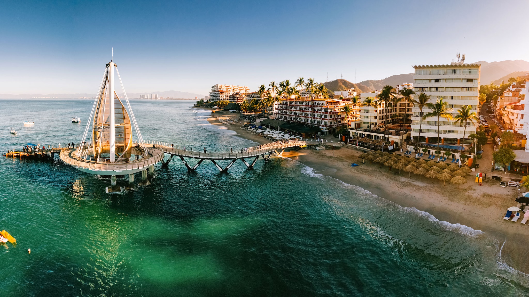 Ein runder Holzsteg im türkisgrünen Meer an einem Sandstrand mit aufgereihten Hotels.