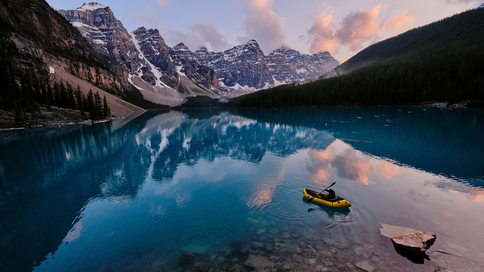 Eine Person in einem Kajak paddelt auf einsamem Gewässer in Kanada, umgeben von schroffen Gipfeln und Tannenwäldern.