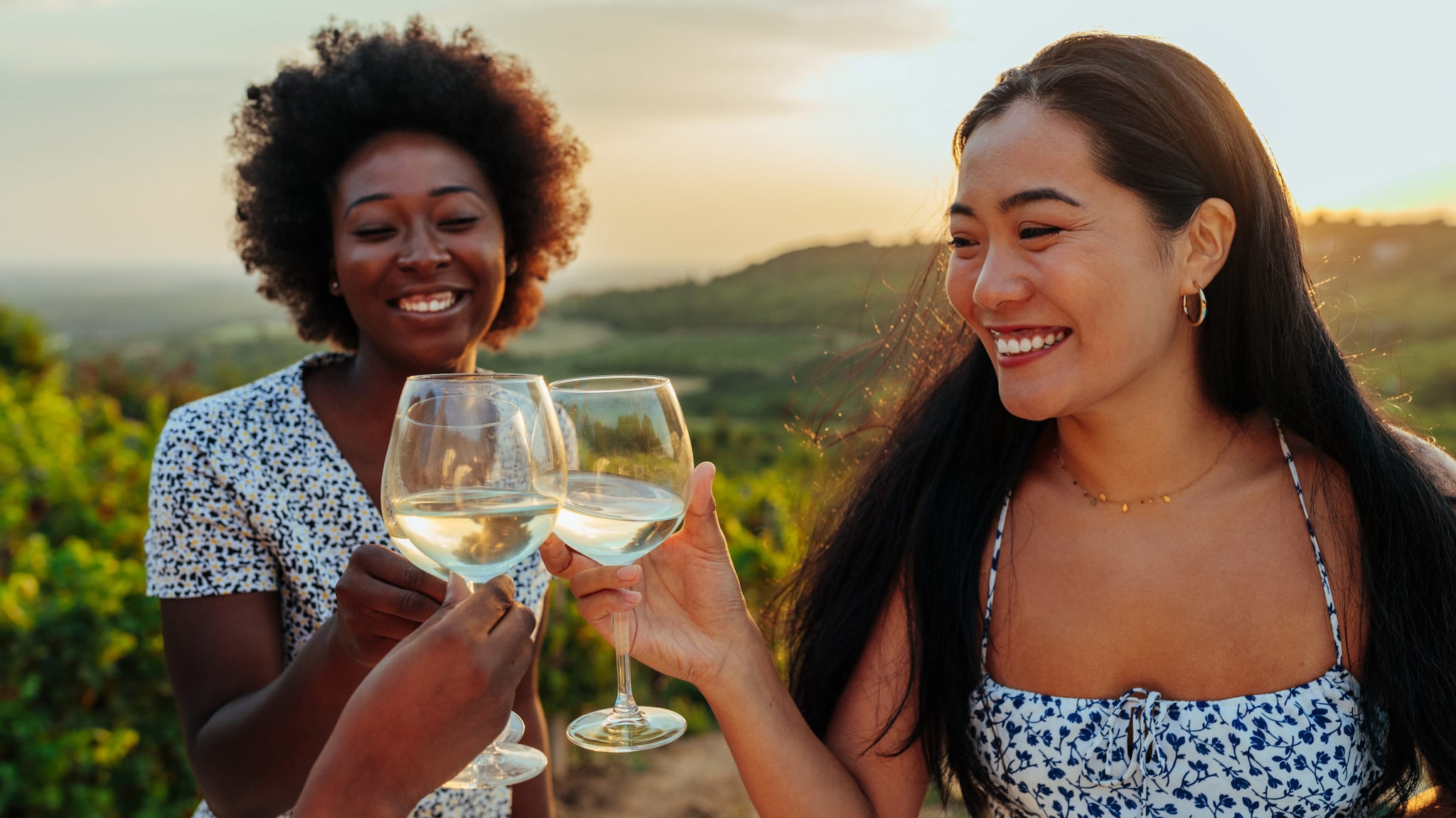 Zwei Frauen stehen in grünen Weingärten und prosten sich mit Weißwein zu.