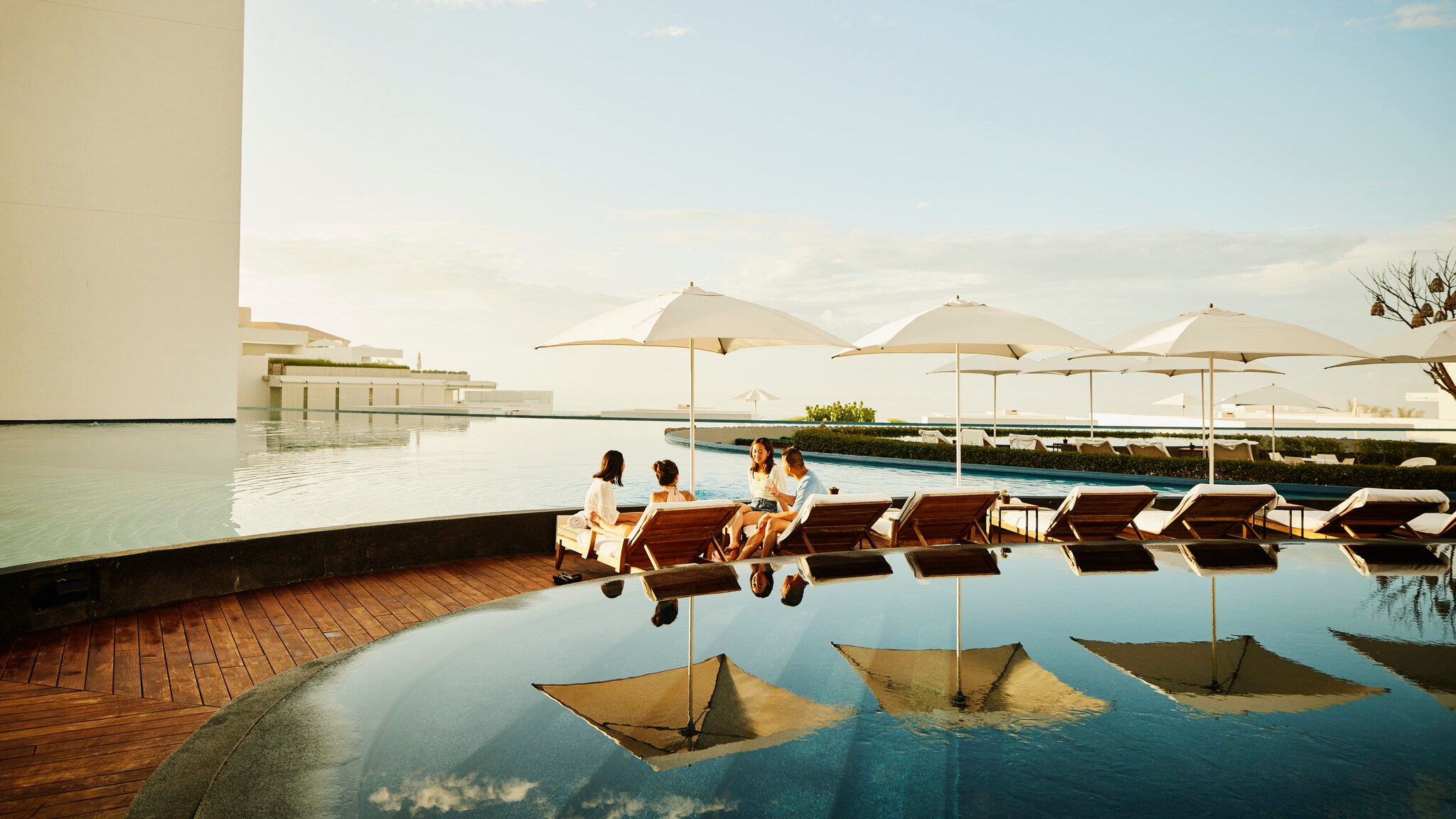 Vier Menschen sitzen unter einem Sonnenschirm, umgeben von einer großen Poollandschaft und der Weite des Himmels.
