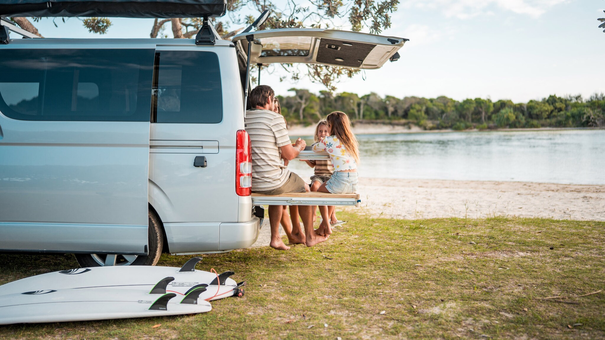 Eine Familie mit zwei Kindern sitzen bei schönem Sommerwetter auf der Ladefläche eines Vans und unterhalten sich.