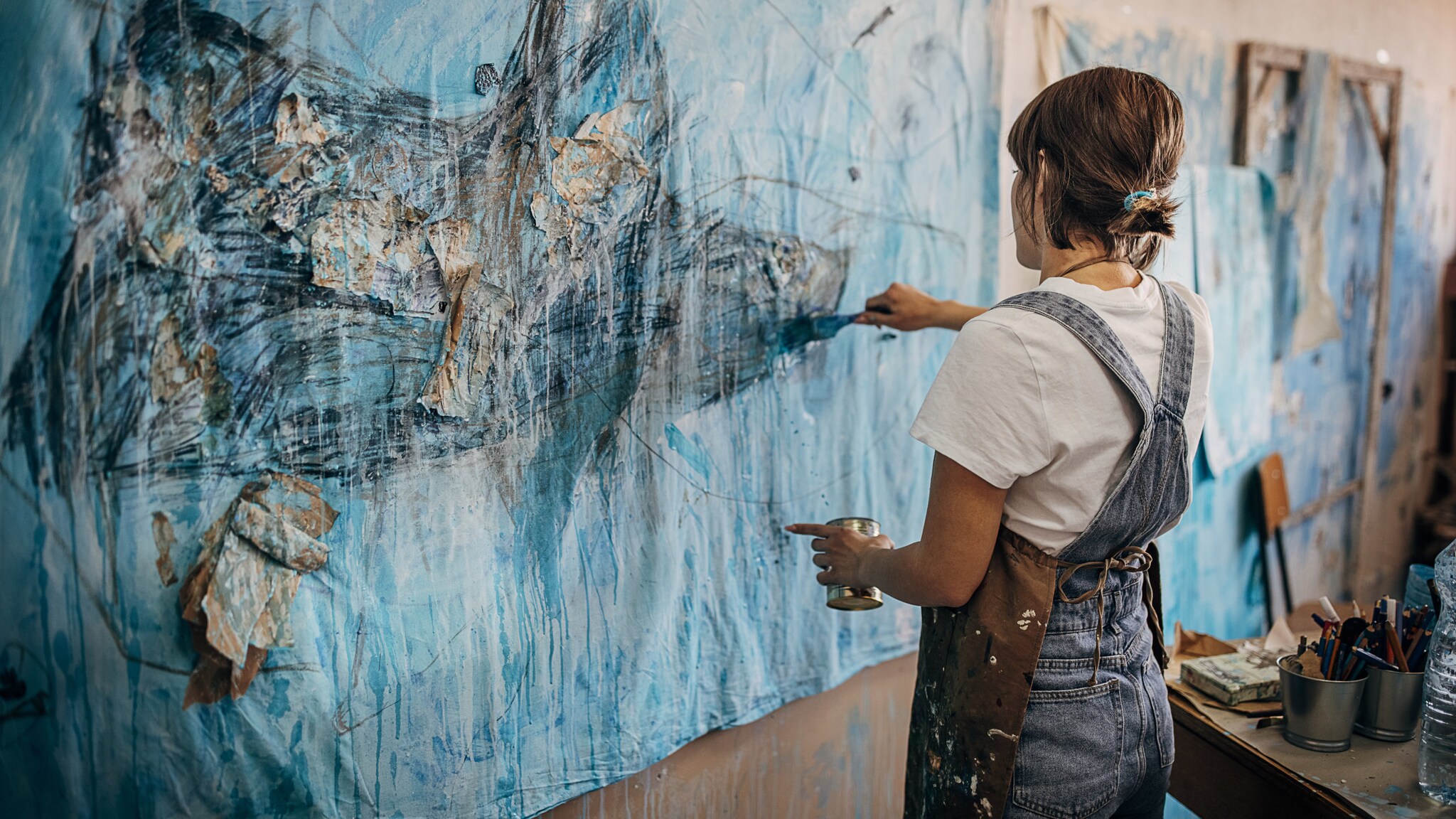 Eine Frau in Jeans-Latzhose steht malend vor einem großen abstrakten Bild.