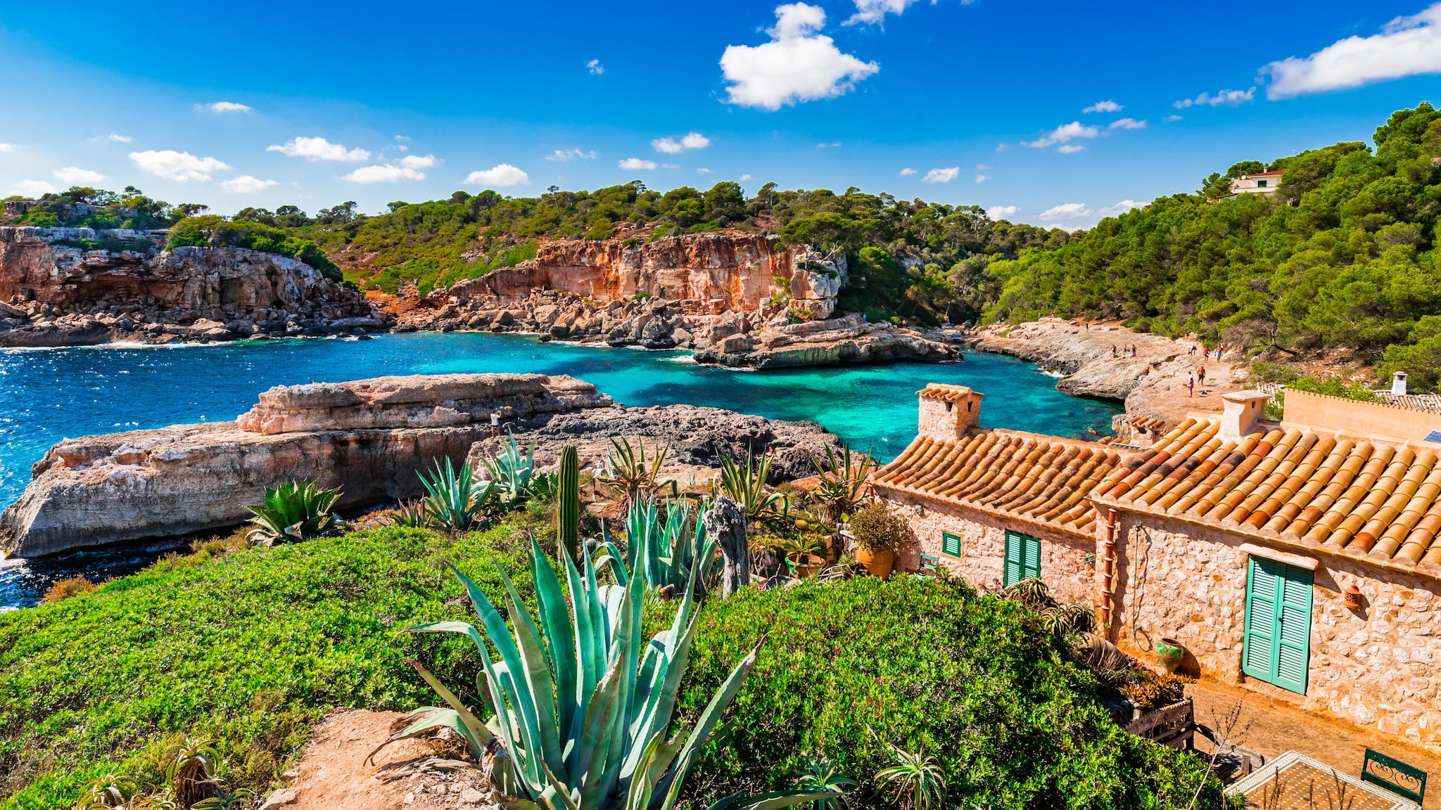 Küstenabschnitt Mallorcas mit üppig-grüner Vegetation, blauem Wasser, schroffen Felsen und einer klassischen Finca.