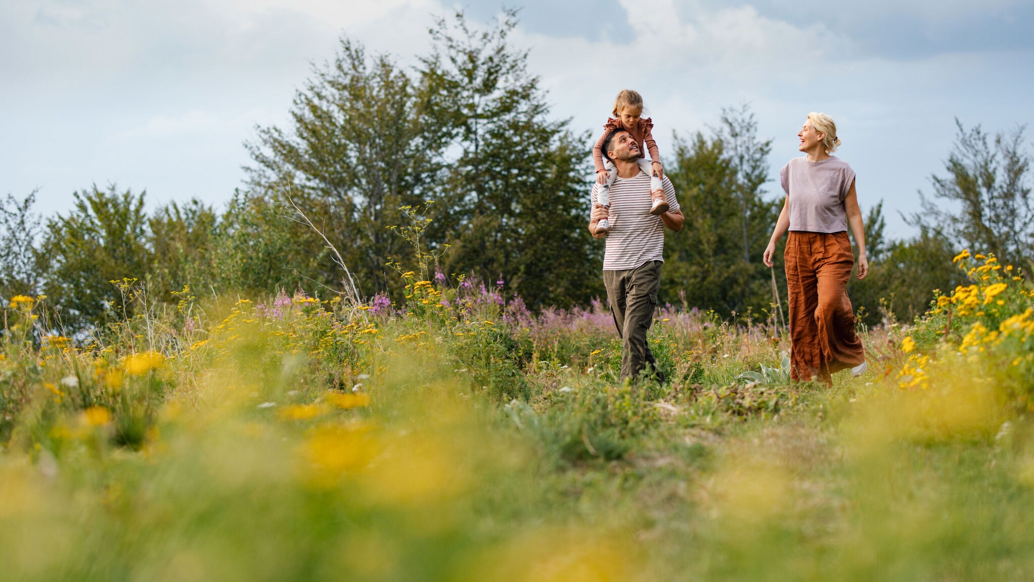 Junge Familie spaziert über eine Naturlandschaft mit bunten Wildblumen, der Vater trägt seine Tochter auf den Schultern.