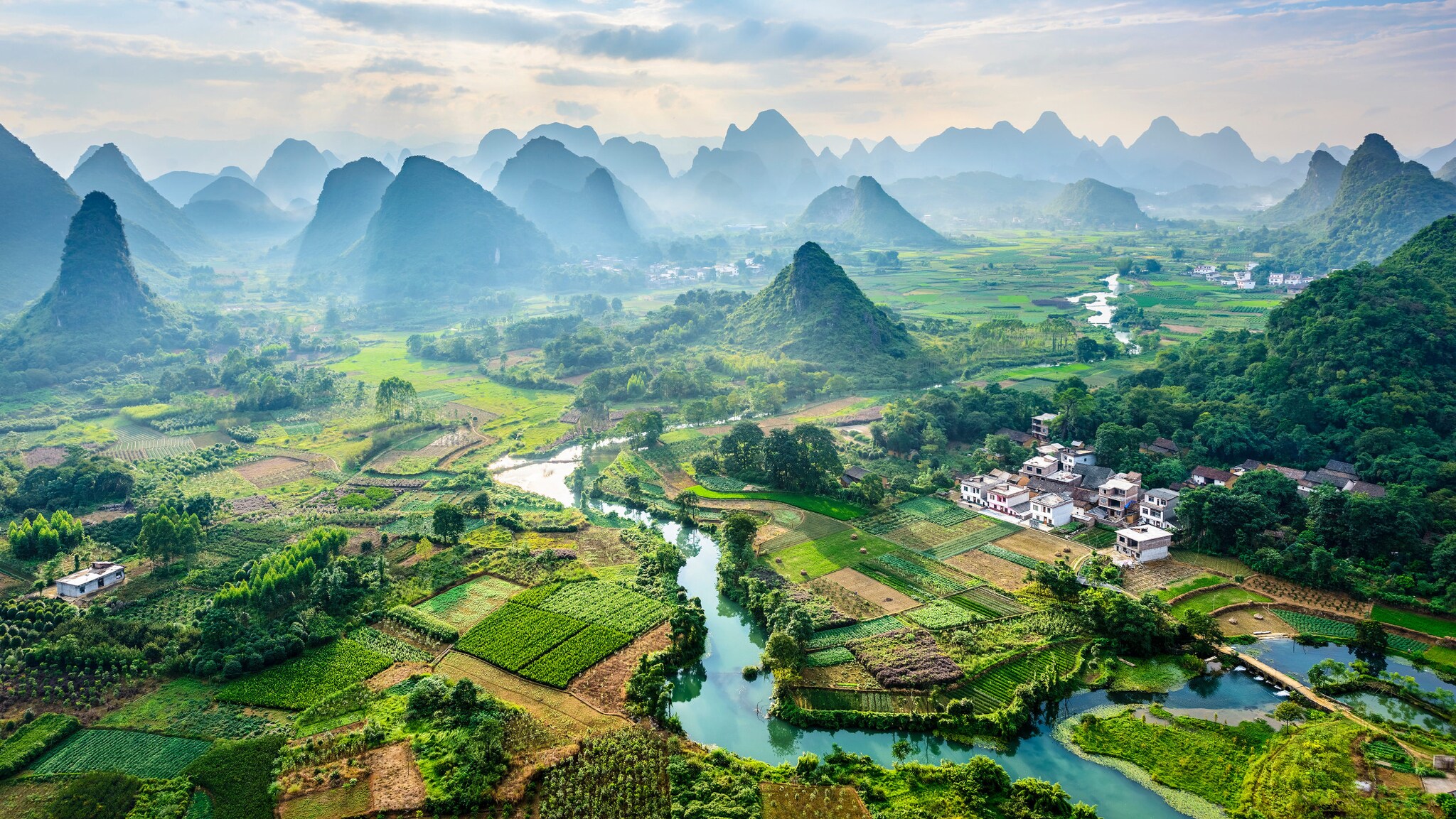 Ostasiatische Landschaft mit satt-grünen Feldern, türkisfarbenem Fluss und, leicht in Nebel gelegenen Hügeln im Hintergrund.