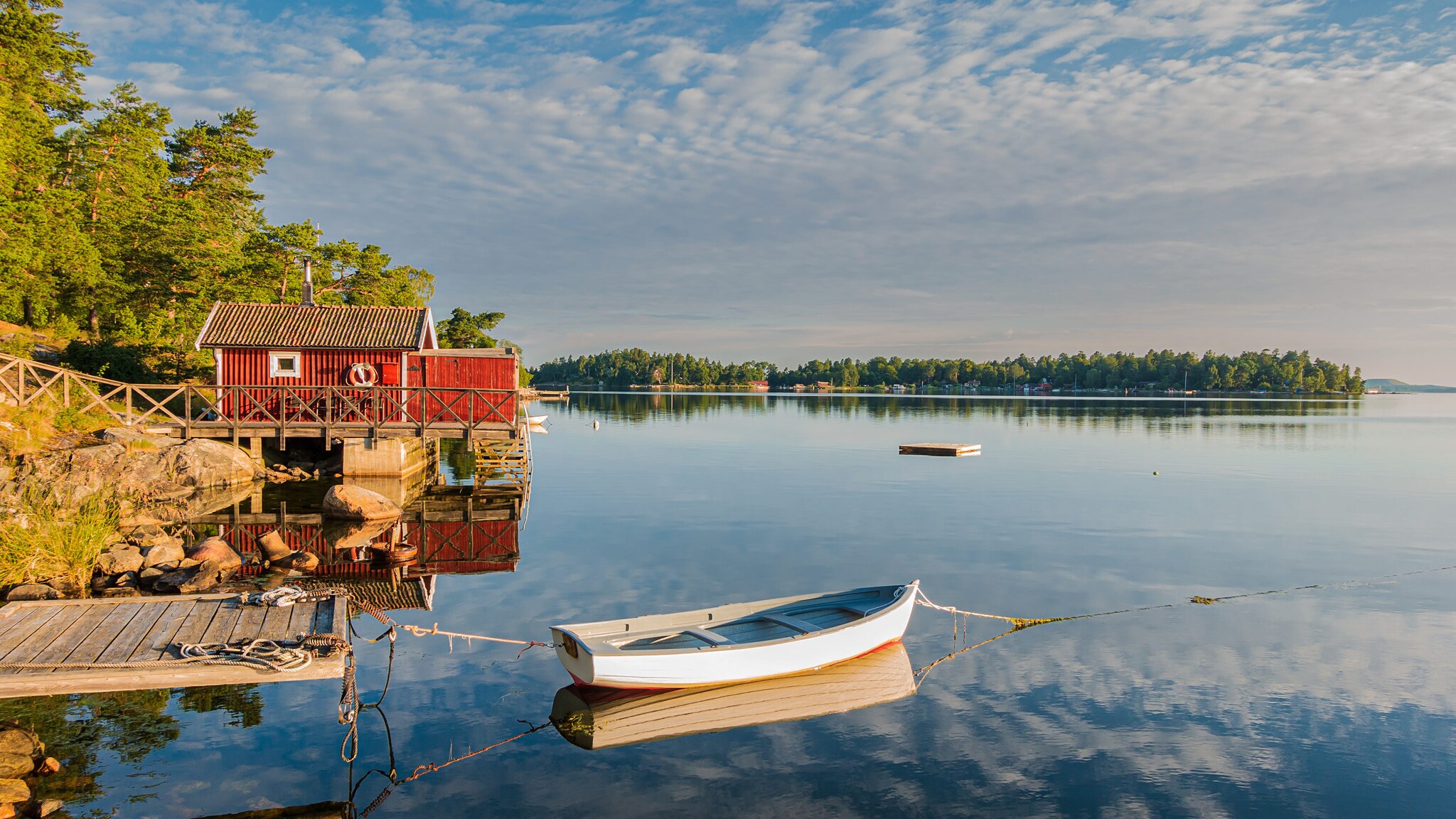 Ein kleines weißes Ruderboot auf einem See, befestigt an einem Steg, dahinter eine rote Holzhütte und grüne Wälder.