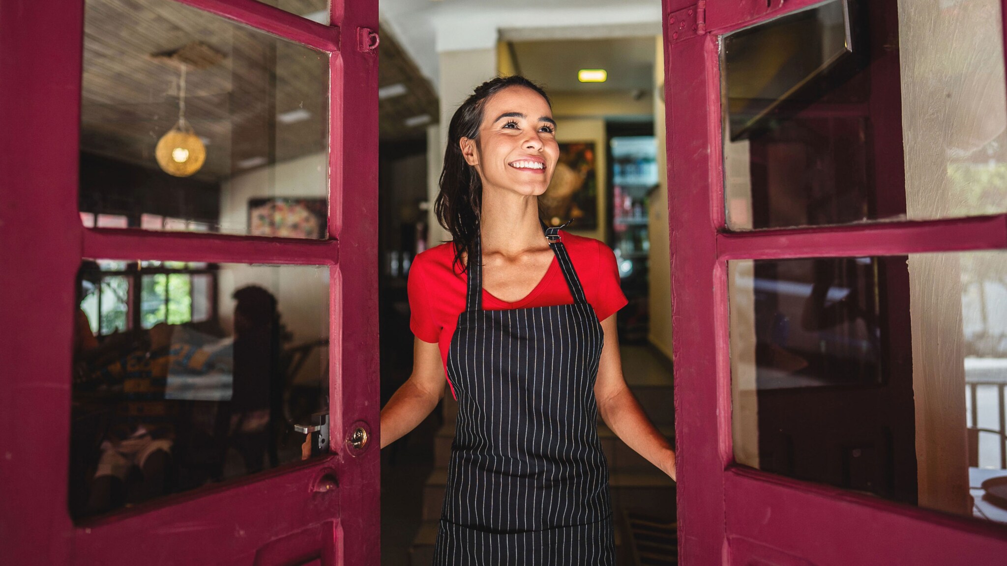 Eine lächelnde Frau mit Pferdeschwanz und dunkler Schürze öffnet eine rote Ladentür mit Glaselementen.