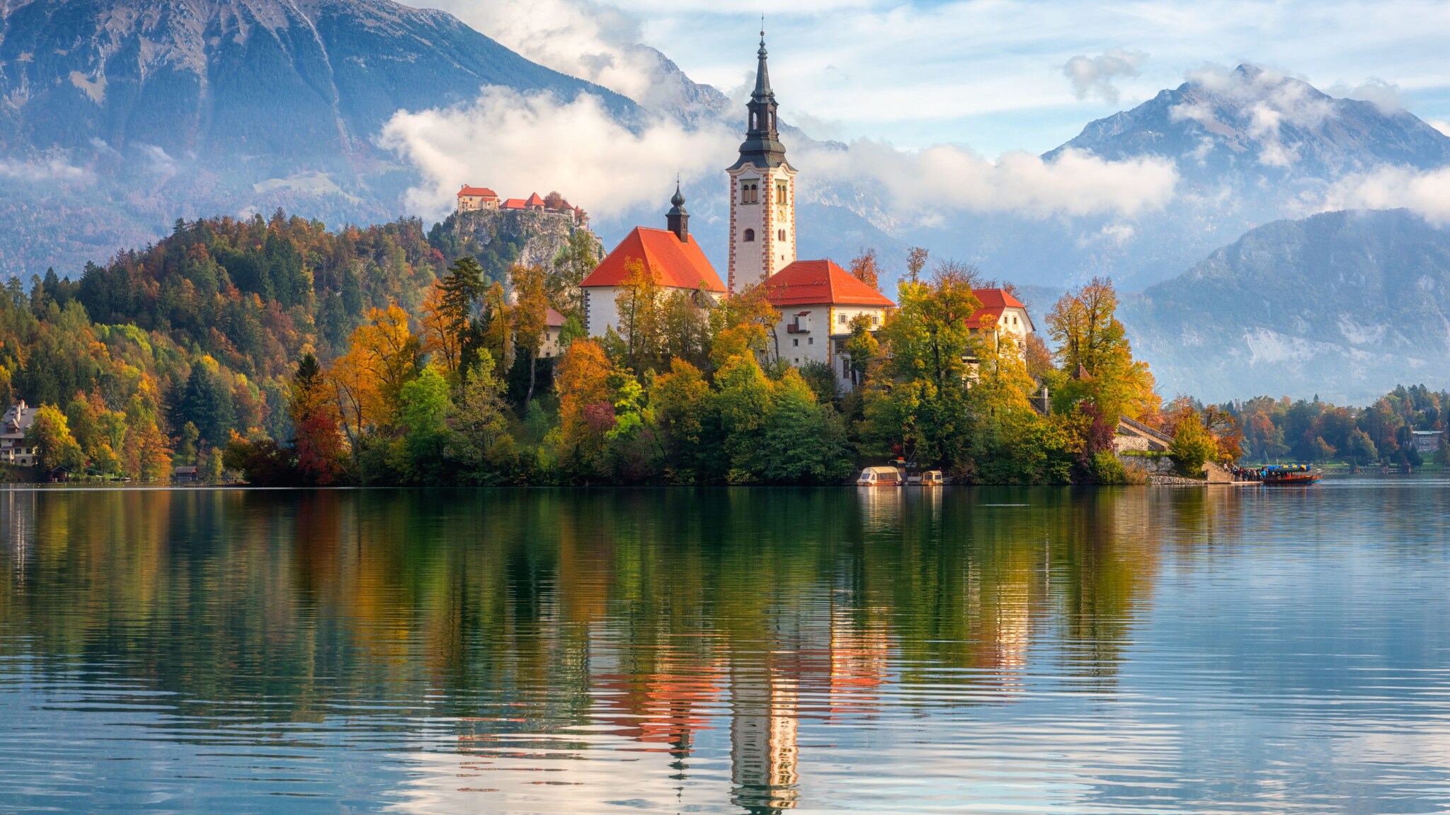Blick auf die malerische Kulisse des Bleder Sees an einem Herbsttag, mit Bleder Burg, Kirche und wolkenverhangenen Bergen im Hintergrund.