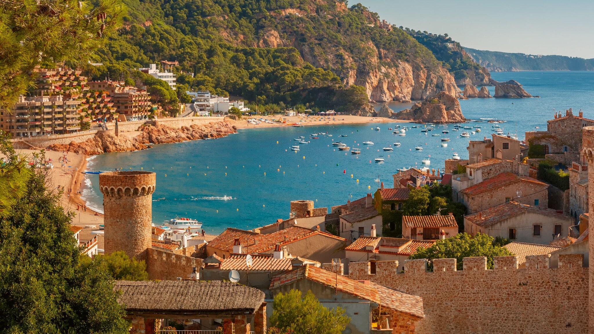 Bucht in Spanien mit goldenem Strand, Boote auf dem blauen Wasser, grün bewachsenen Felsen und traditionellen alten Steinhäusern im Vordergrund.
