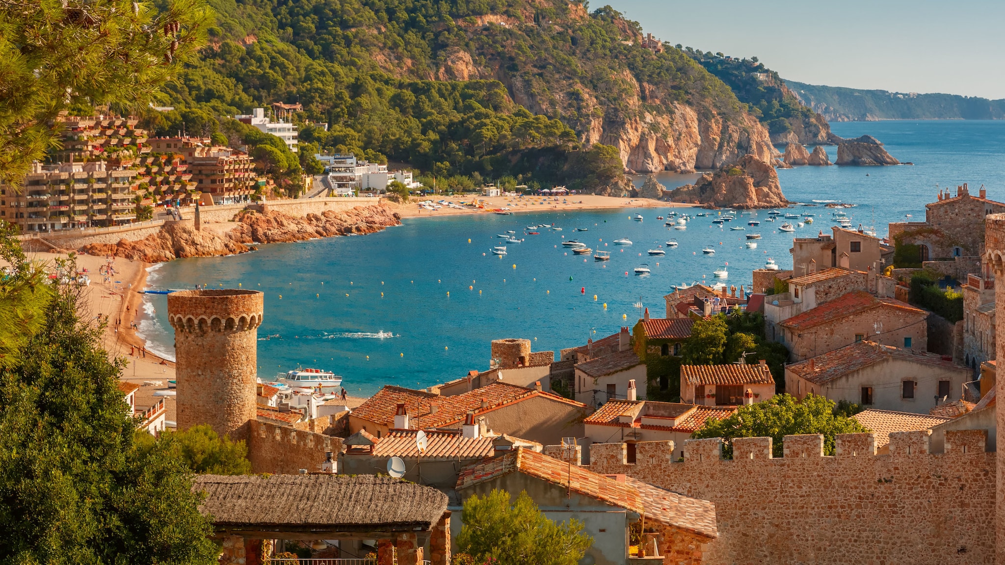 Bucht in Spanien mit goldenem Strand, Boote auf dem blauen Wasser, grün bewachsenen Felsen und traditionellen alten Steinhäusern im Vordergrund.