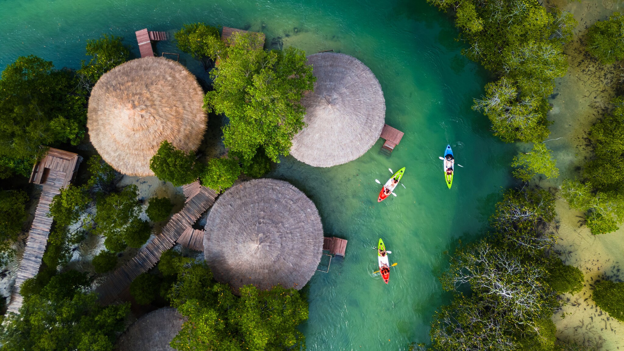 Blick von oben auf bunte Kajaks, die einen türkisfarbenen Fluss entlangfahren, der sich durch grüne Mangrovenlandschaft und an drei traditionellen Hütten mit Strohdach vorbei schlängelt.