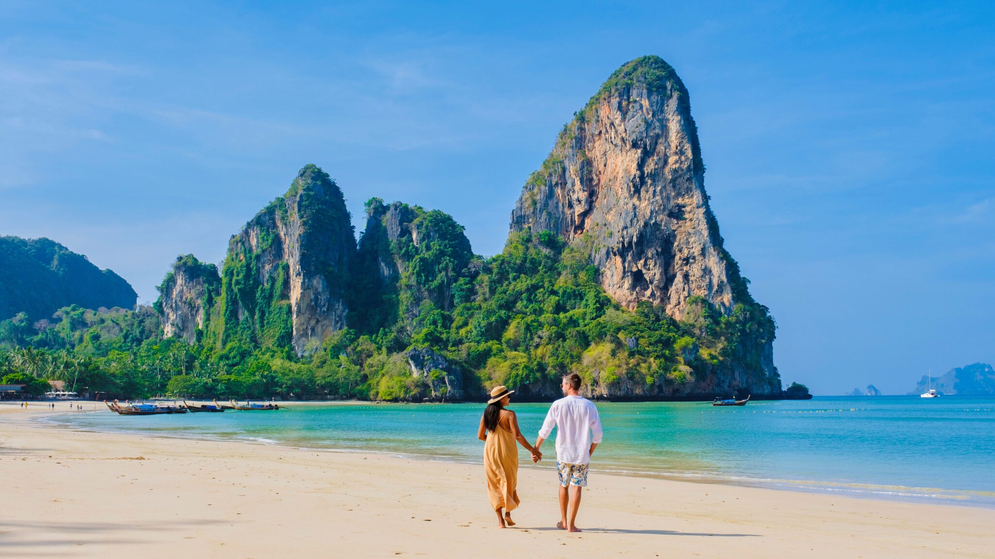 Ein Paar spaziert Hand in Hand am Strand entlang, im Hintergrund liegen traditionell-thailändische Boote im Wasser und schroffe grün bewachsene Felsen ragen empor.