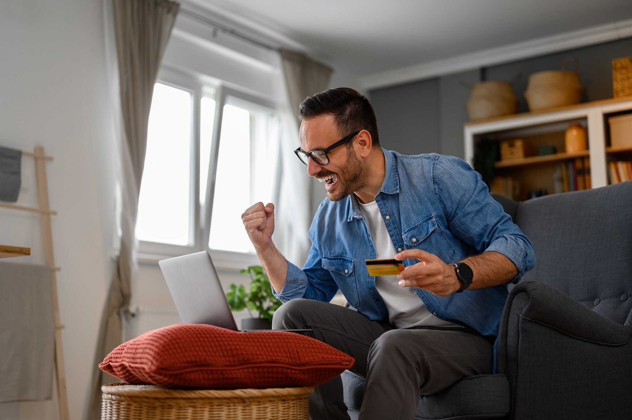 Moon-Safari-iStock-1589525413 Ein Mann sitzt auf einem Sofa vor einem aufgeklappten Laptop, der auf einem Hocker steht. In einer Hand hält er eine Geldkarte, die andere ballt er zu einer Faust.
