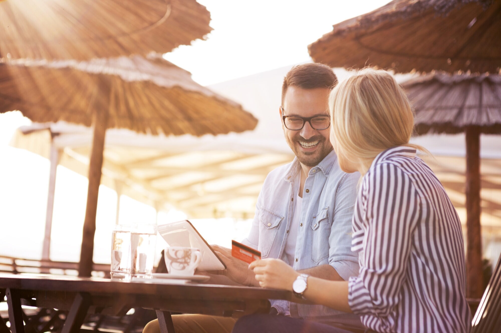 Junges Paar sitzt lachend unter Strandschirmen mit Tablet und Kreditkarte. Junges Paar sitzt lachend unter Strandschirmen mit Tablet und Kreditkarte.
