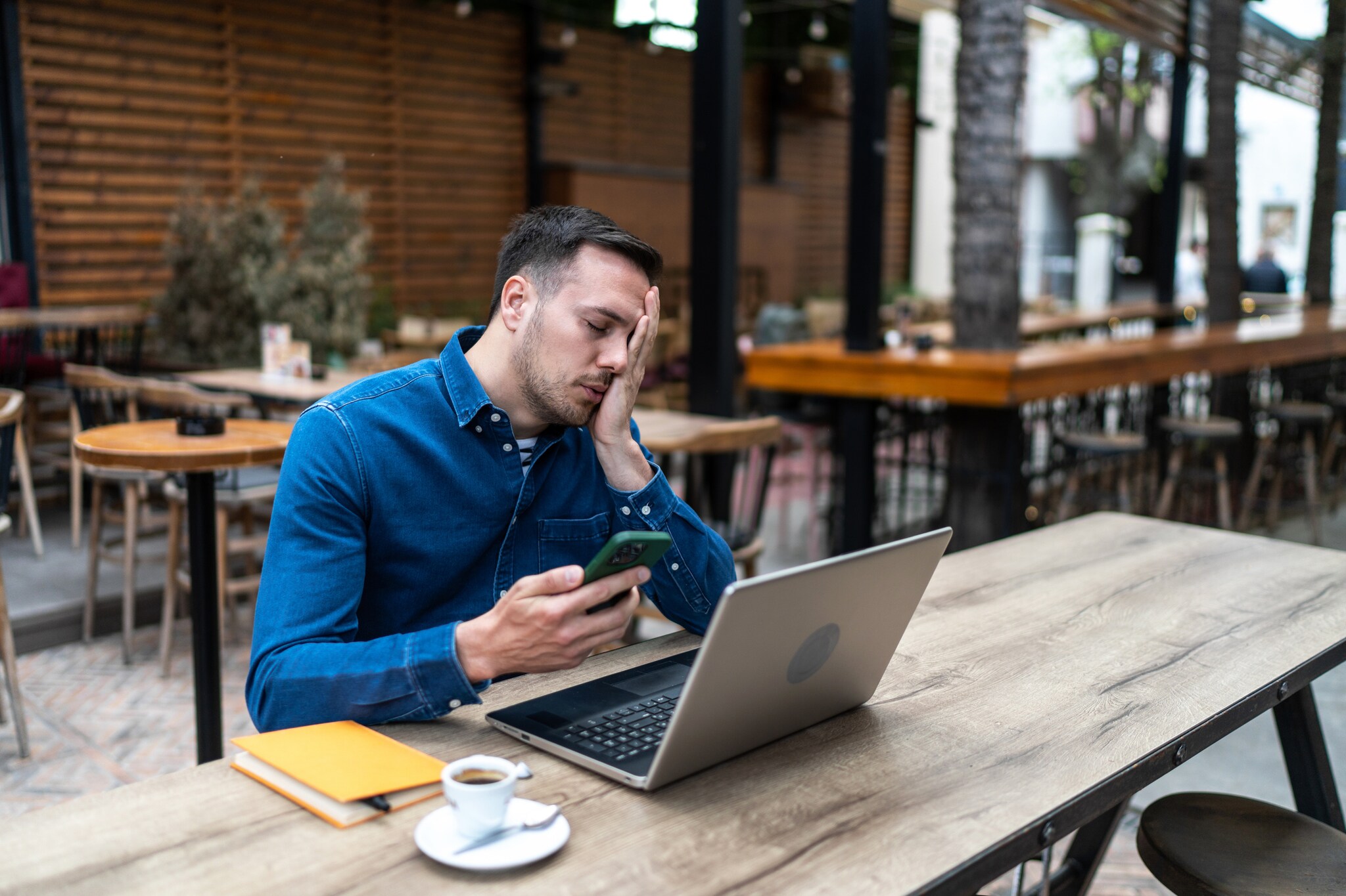Ein Mann sitzt an einem Tisch vor einem Laptop und hält ein Smartphone in der Hand. Er hat die Augen geschlossen und legt eine Hand über sein Gesicht.