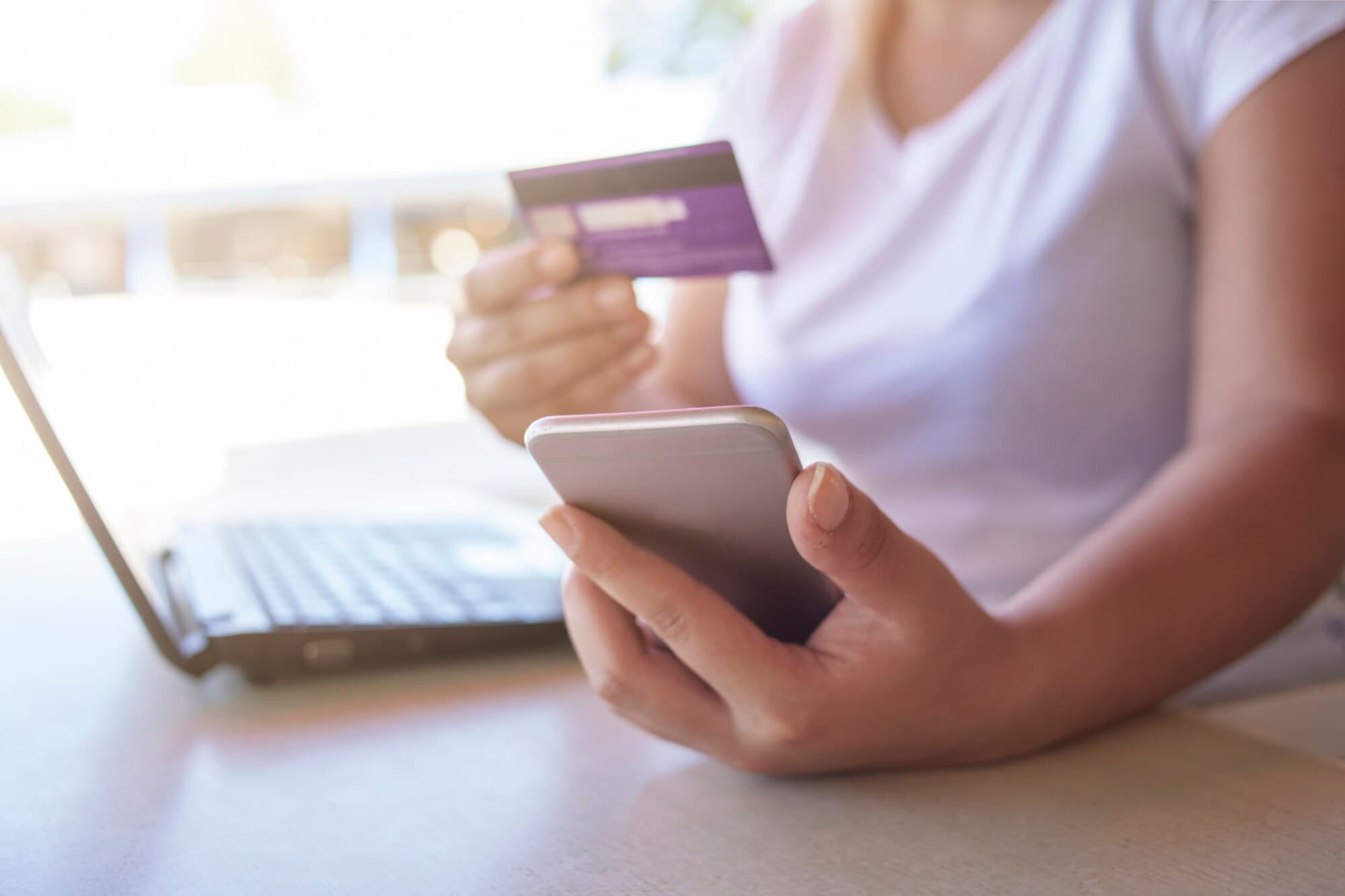 Eine Person sitzt am Tisch vor einem aufgeklappten Notebook. In Händen hält sie Smartphone und Kreditkarte. Eine Person sitzt am Tisch vor einem aufgeklappten Notebook. In Händen hält sie Smartphone und Kreditkarte.