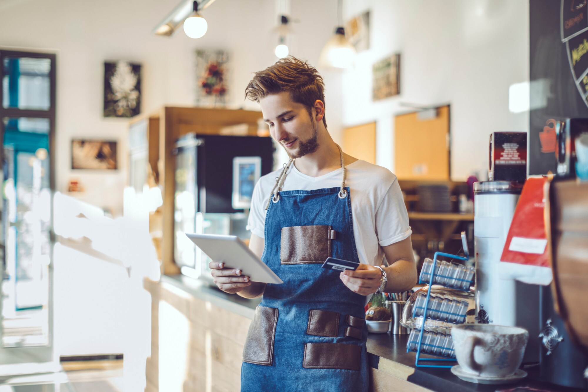 Ein junger Barista steht in einem Café, er hat eine Schürze an und hält in der einen Hand ein Tablet und in der anderen Hand eine Kreditkarte