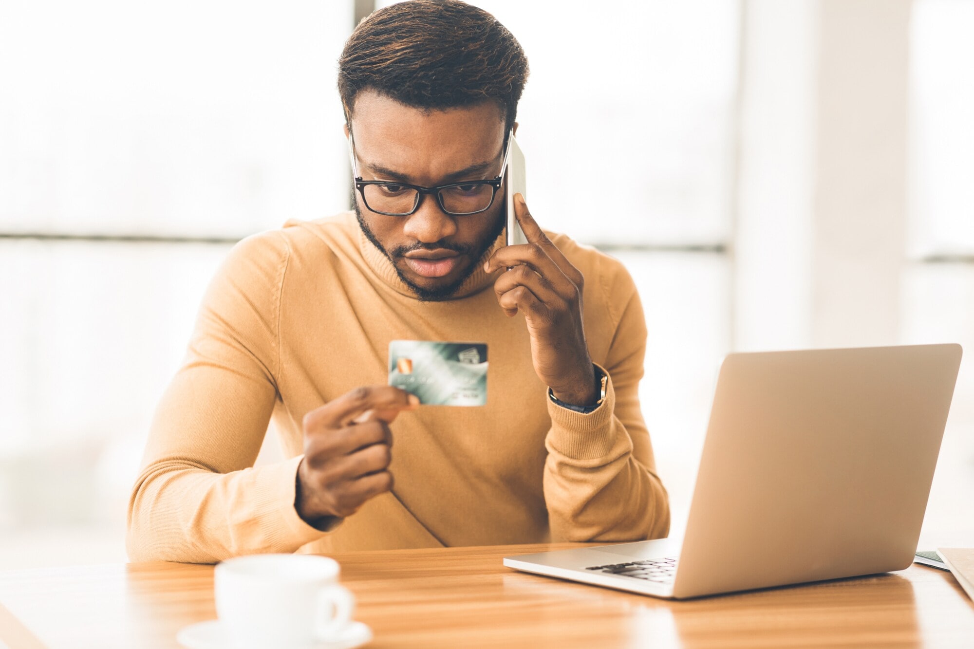 Eine Person sitzt telefonierend vor einem geöffneten Laptop am Tisch und blickt auf seine Bankkarte in der Hand. Eine Person sitzt telefonierend vor einem geöffneten Laptop am Tisch und blickt auf seine Bankkarte in der Hand.