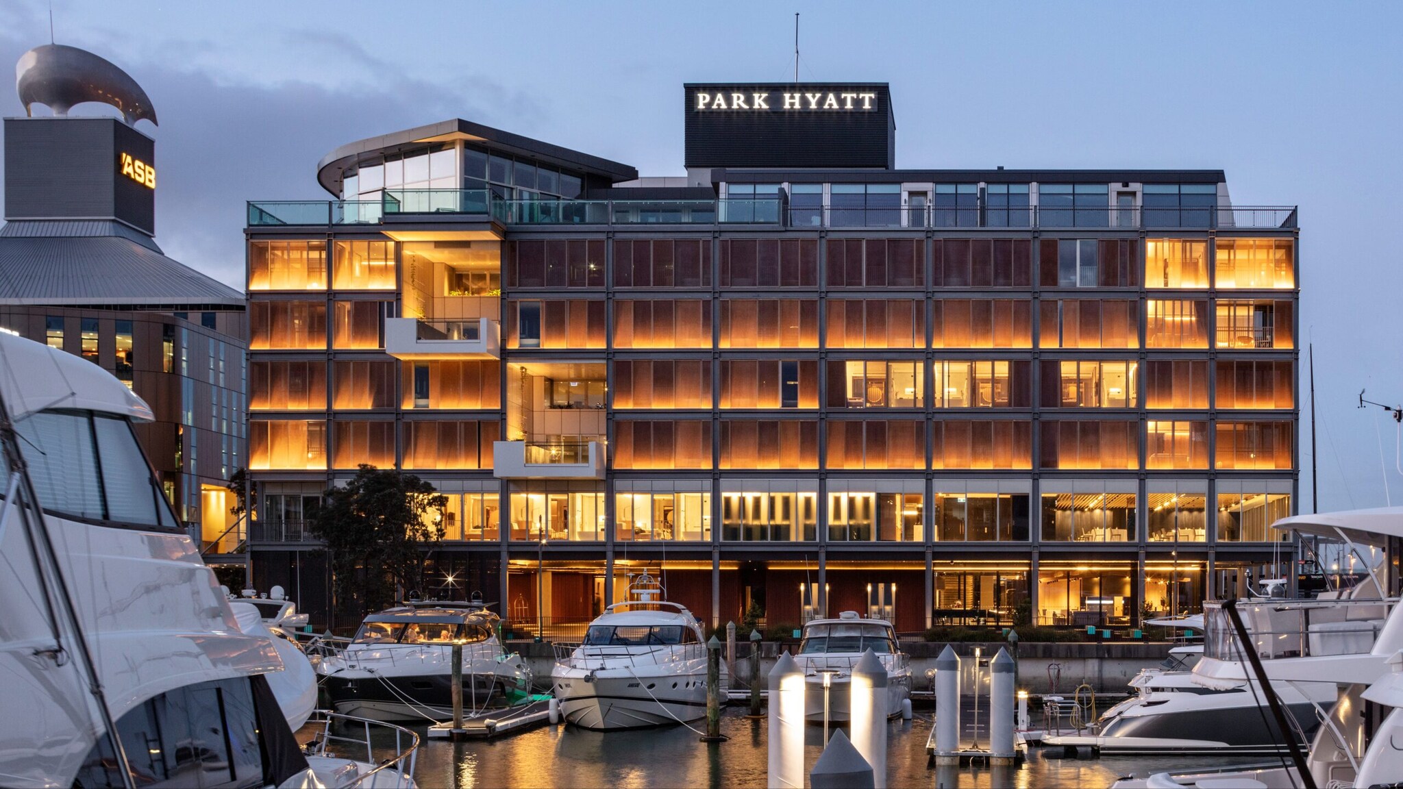 Das warme Licht des Park Hyatt Hotels spiegelt sich im Hafenbecken von Auckland, während die weißen Yachten direkt davor vor Anker liegen.