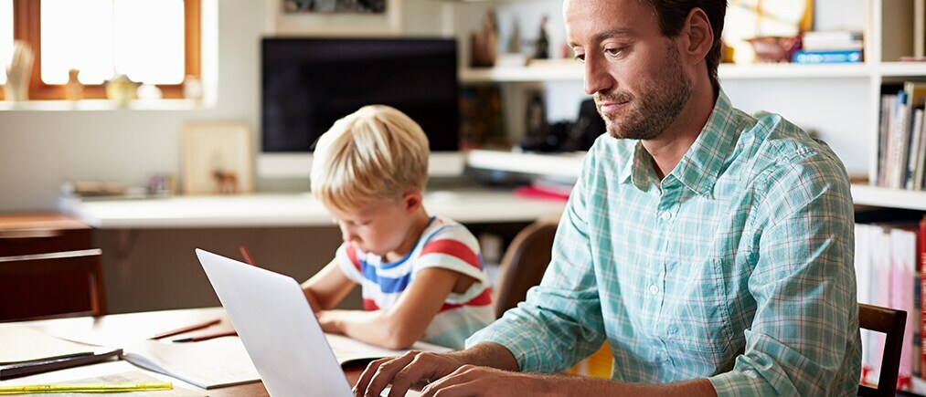 Blogbild: Person arbeitet am laptop während ein Kind daneben Hausaufgaben macht
