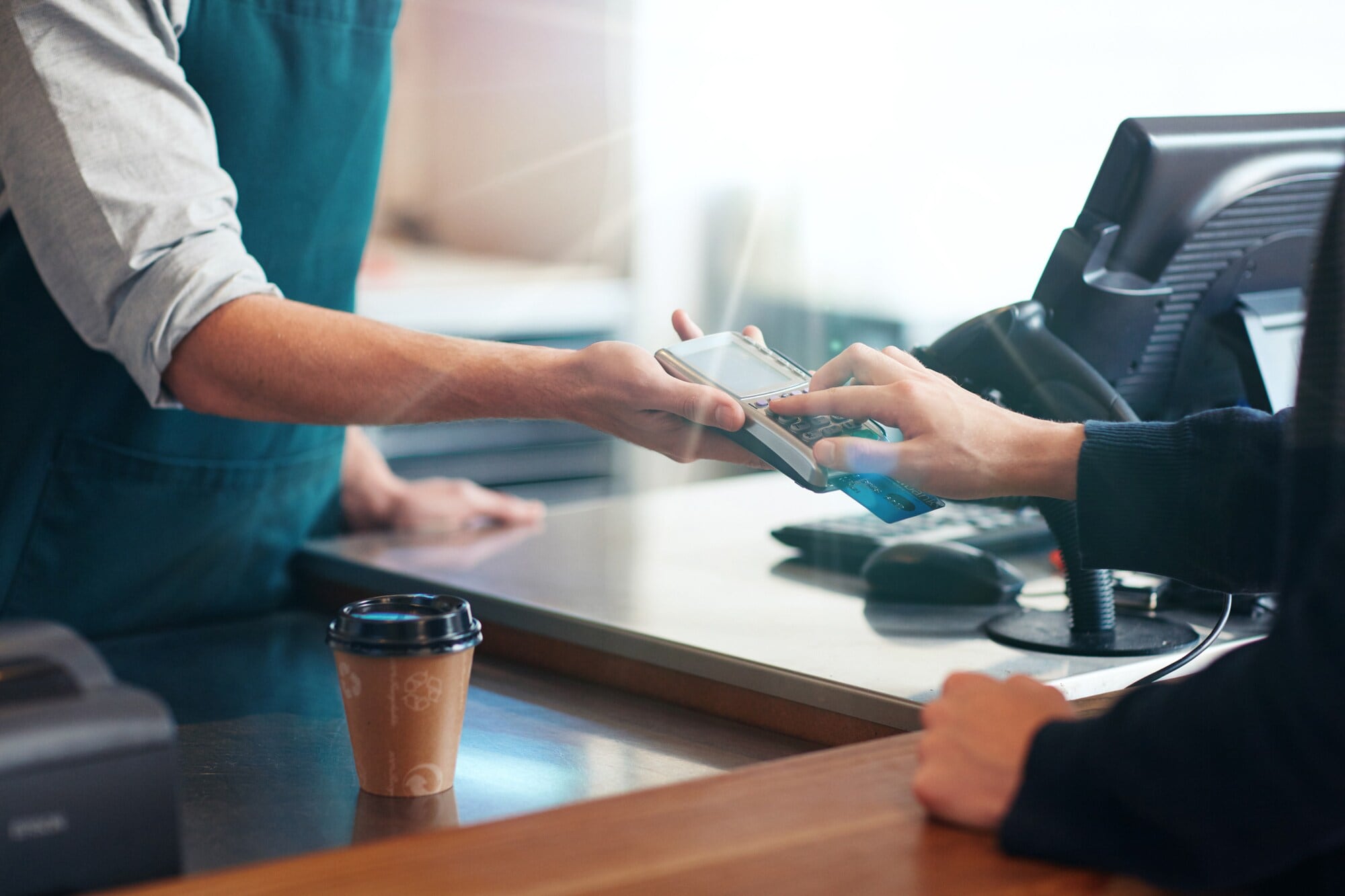 Eine Person, die über einen Tresen, auf dem ein Kaffeebecher steht, Zahlen in ein Terminal eingibt, das eine Person auf der anderen Seite des Tresens hinhält Eine Person, die über einen Tresen, auf dem ein Kaffeebecher steht, Zahlen in ein Terminal eingibt, das eine Person auf der anderen Seite des Tresens hinhält