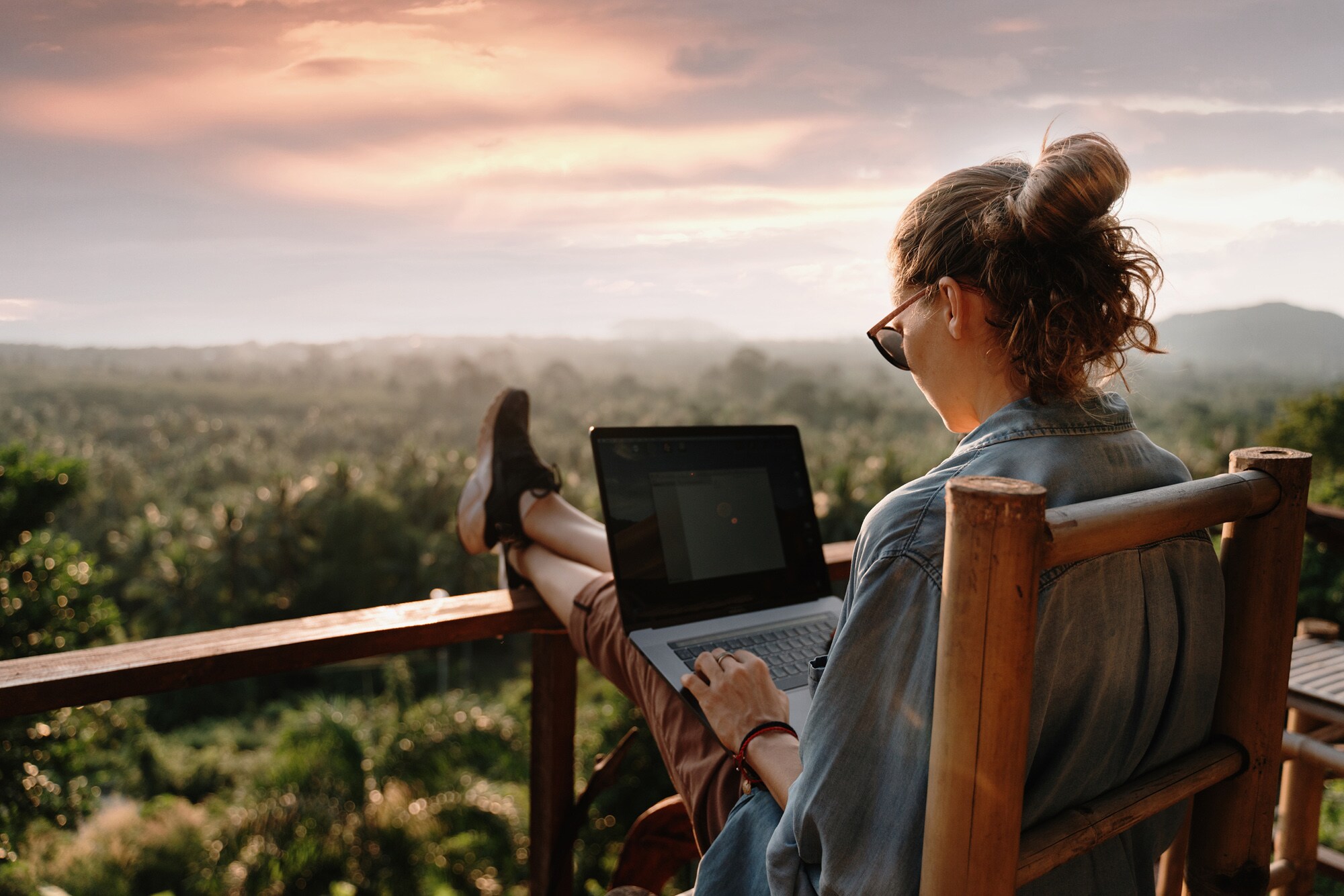 Eine Frau sitzt im Freien auf einer Terrasse, arbeitet an einem Laptop mit Blick ins Grüne.