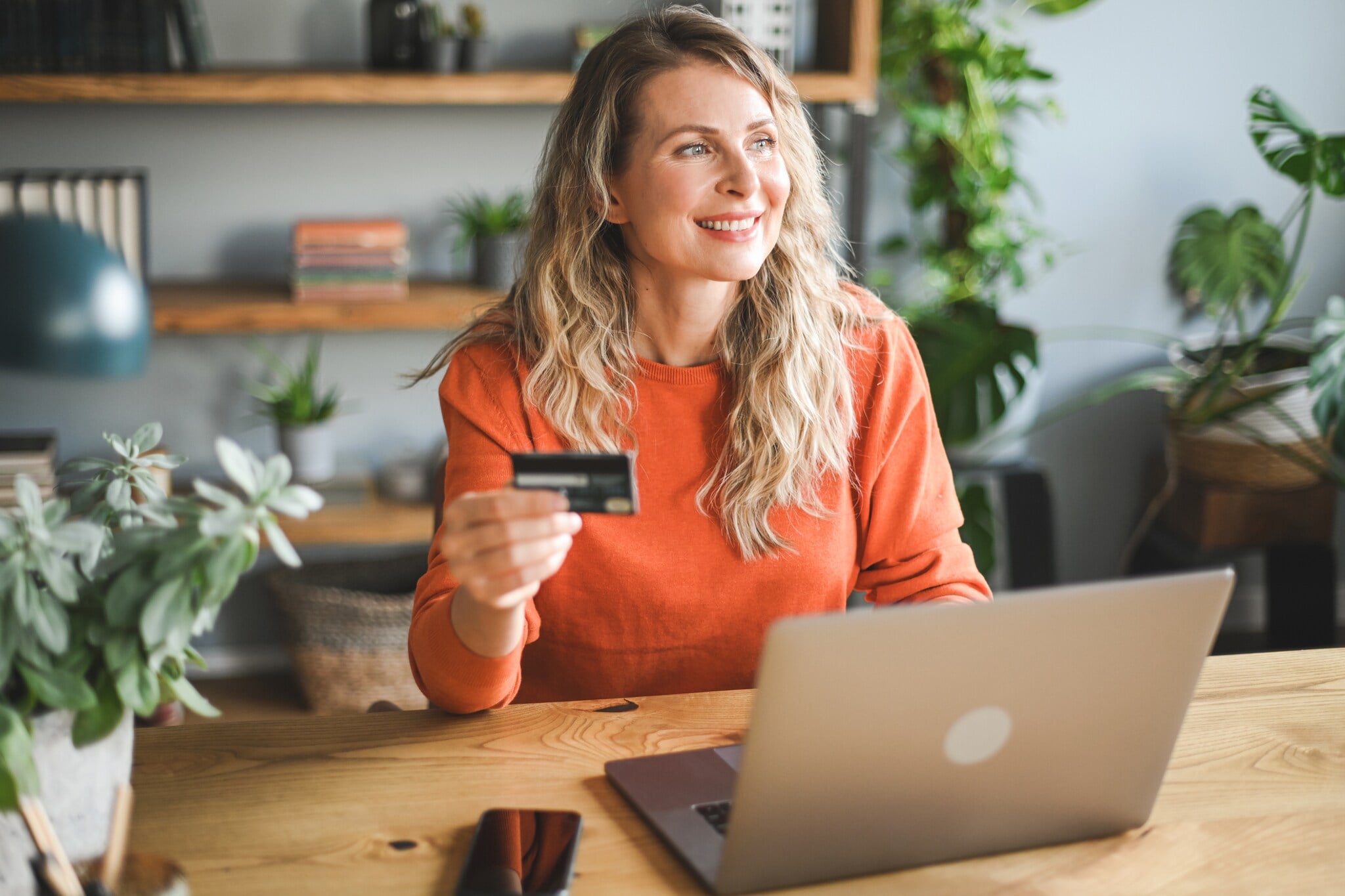 Paypal Eine Frau sitzt lächelnd an einem Tisch. Vor ihr steht ein Laptop und in der Hand hält sie eine Kreditkarte.