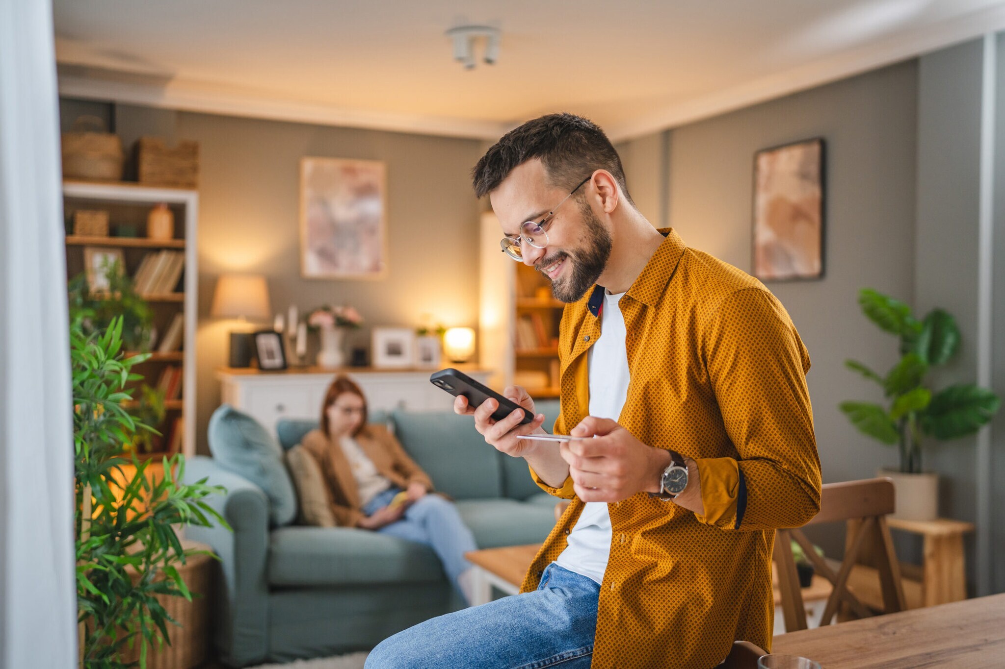 Eine Person sitzt auf einem Tisch und hält eine Kreditkarte sowie ein Smartphone in den Händen. Im Hintergrund sitzt eine weitere Person auf einem Sofa.