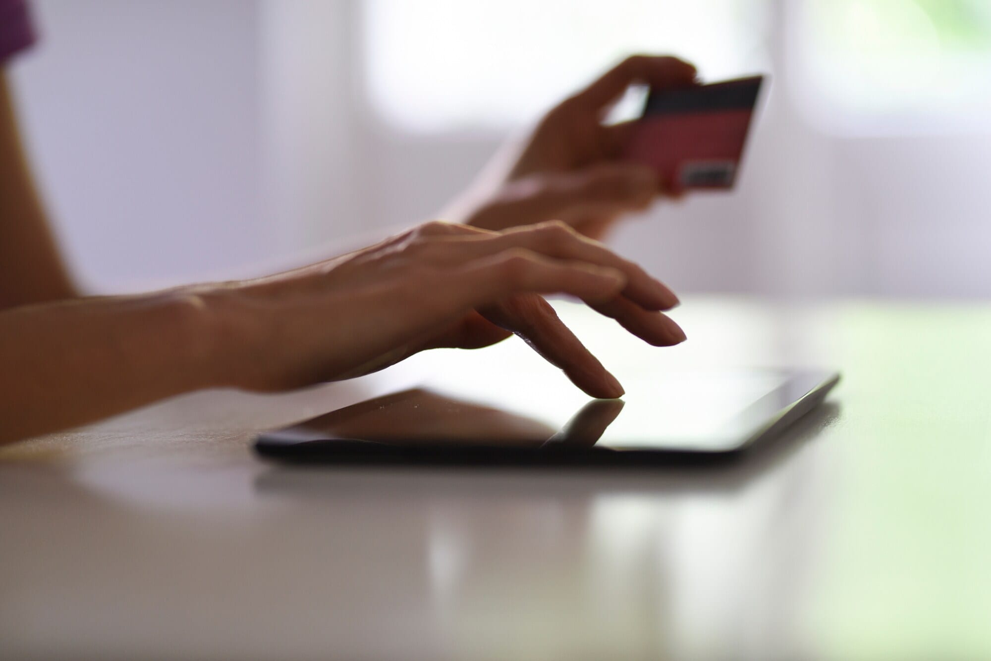 Nahaufnahme einer Hand, die auf einem Tablet tippt. Im Hintergrund ist eine zweite Hand zu erkenne, die eine Bankkarte in der Hand hält. Nahaufnahme einer Hand, die auf einem Tablet tippt. Im Hintergrund ist eine zweite Hand zu erkenne, die eine Bankkarte in der Hand hält.