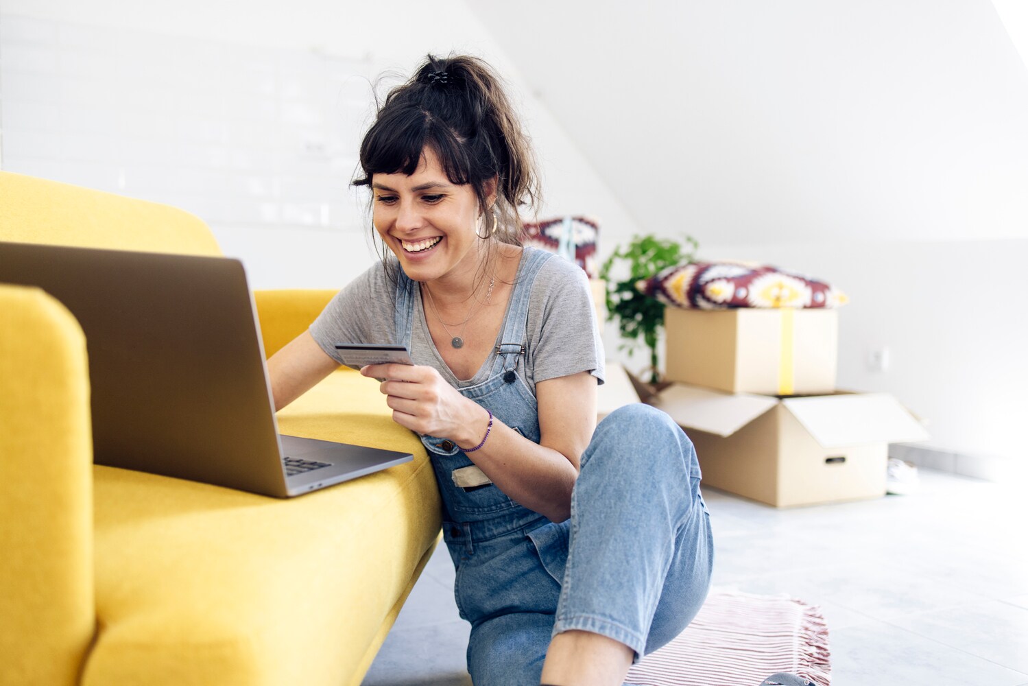 Eine junge Frau mit Kreditkarte in einer Hand vor einem gelben Sofa, auf dem ein Laptop steht