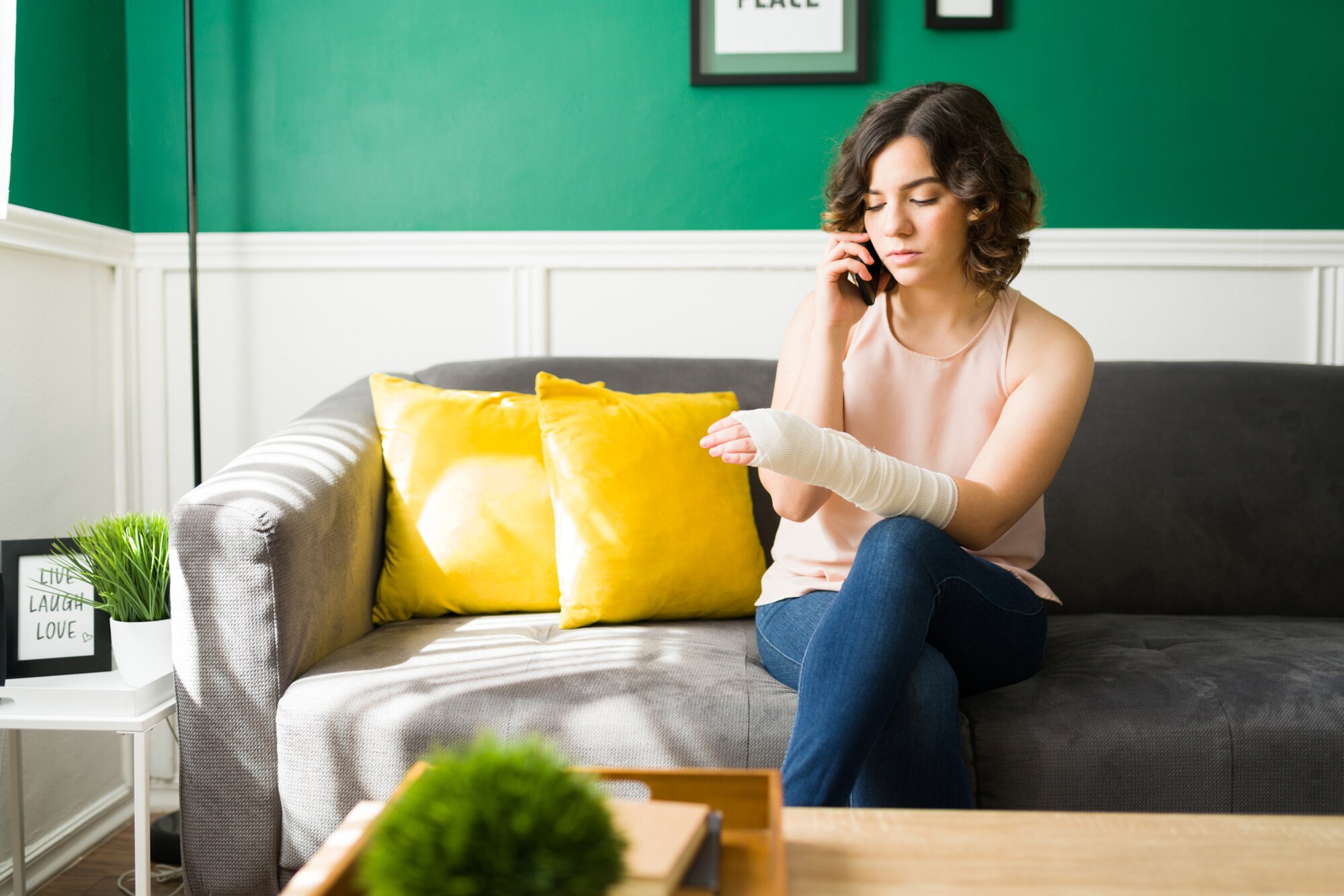 Eine Person sitzt mit einbandagiertem Arm auf einem Sofa und telefoniert.