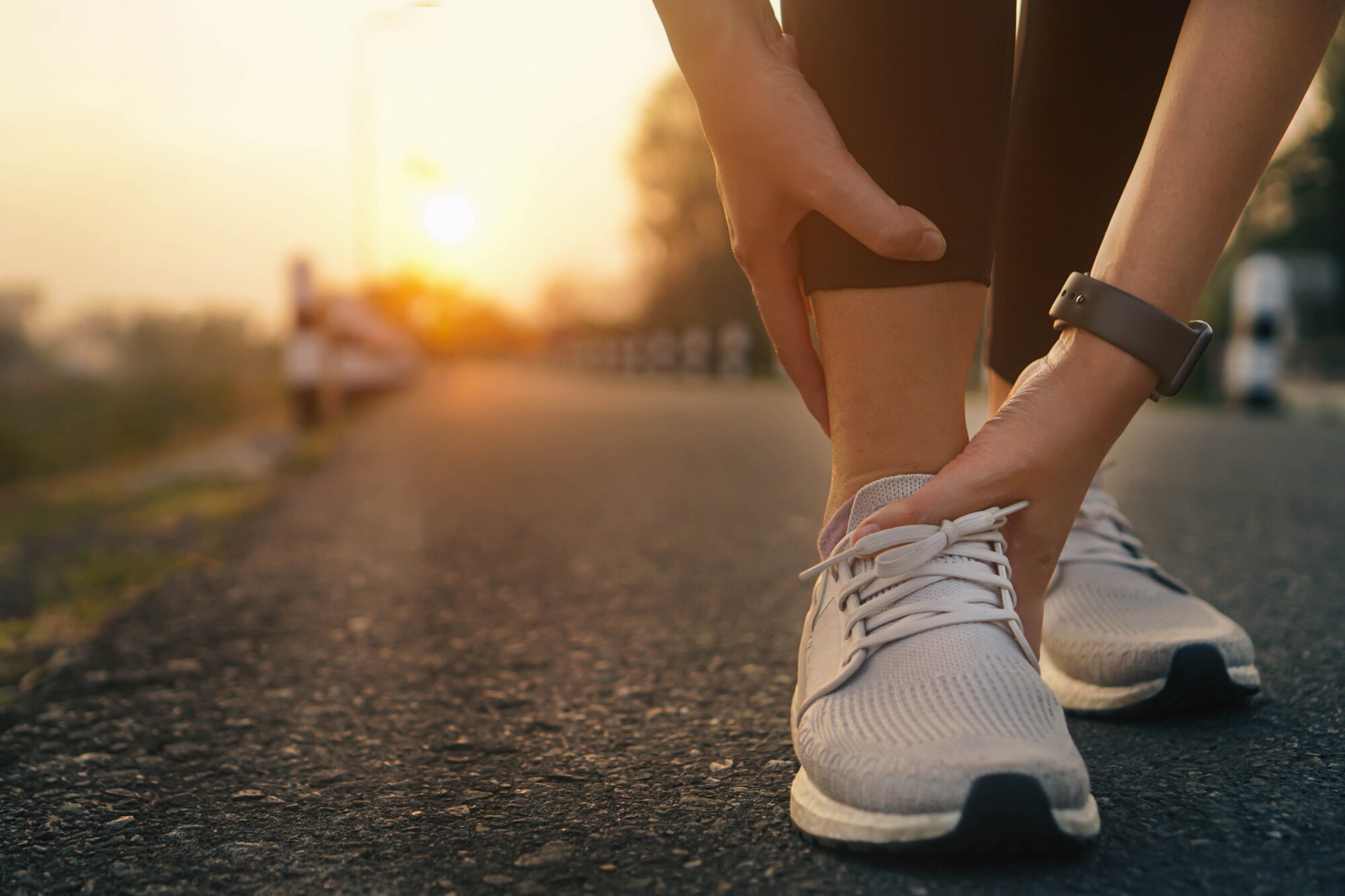 Eine Person in Laufschuhen hockt auf einem asphaltierten Weg und hält sich mit beiden Händen den Knöchel. Eine Person in Laufschuhen hockt auf einem asphaltierten Weg und hält sich mit beiden Händen den Knöchel.