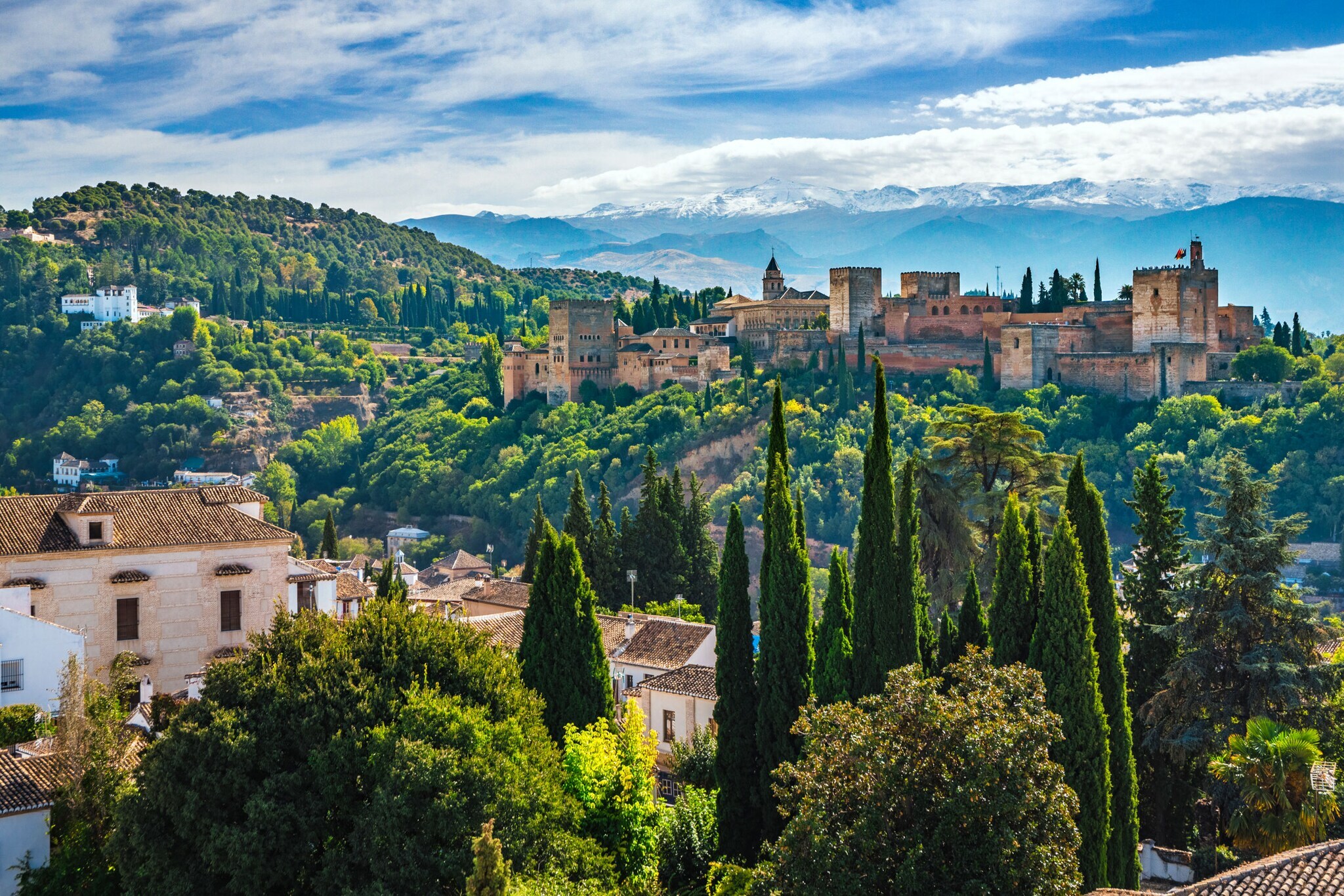 Blick auf einzelne Häuser und die von Wald umgebene Alhambra, im Hintergrund Berge.