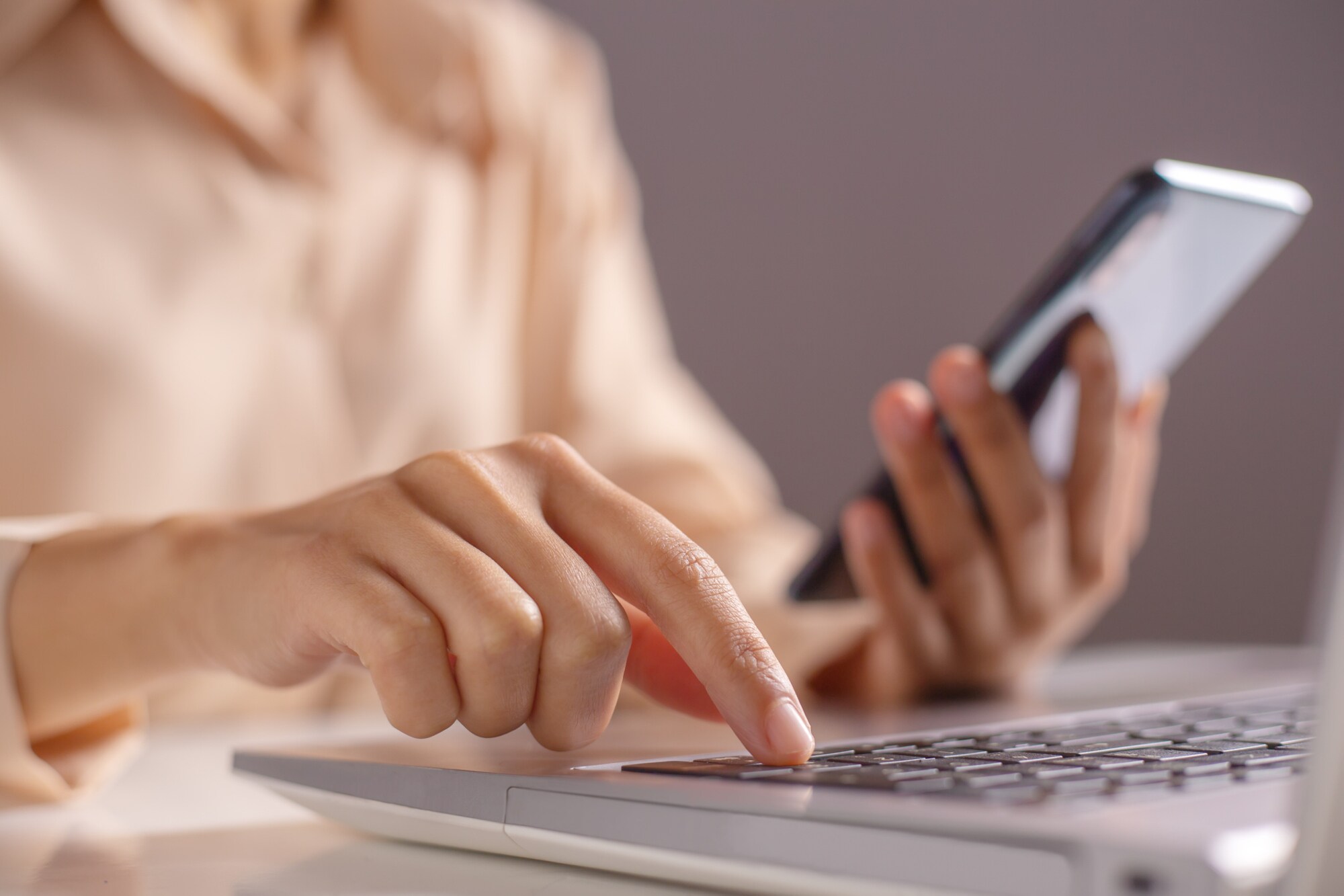 Eine Frau hält ein Handy in der linken Hand und drückt mit der rechten Hand eine Taste auf ihrem Laptop