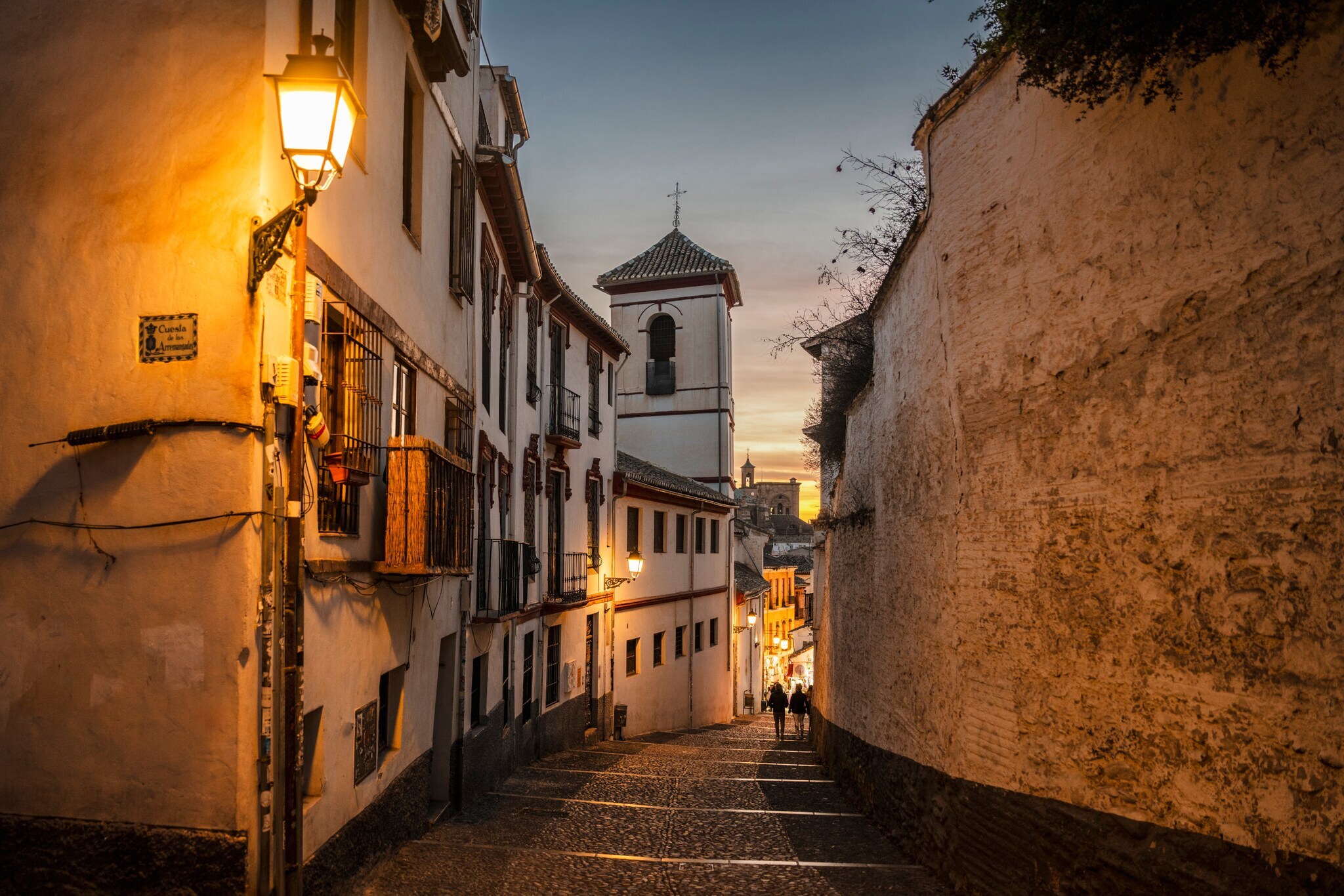 Altstadtgasse zwischen weißen Häusern und einer hohen Mauer in der Abenddämmerung.
