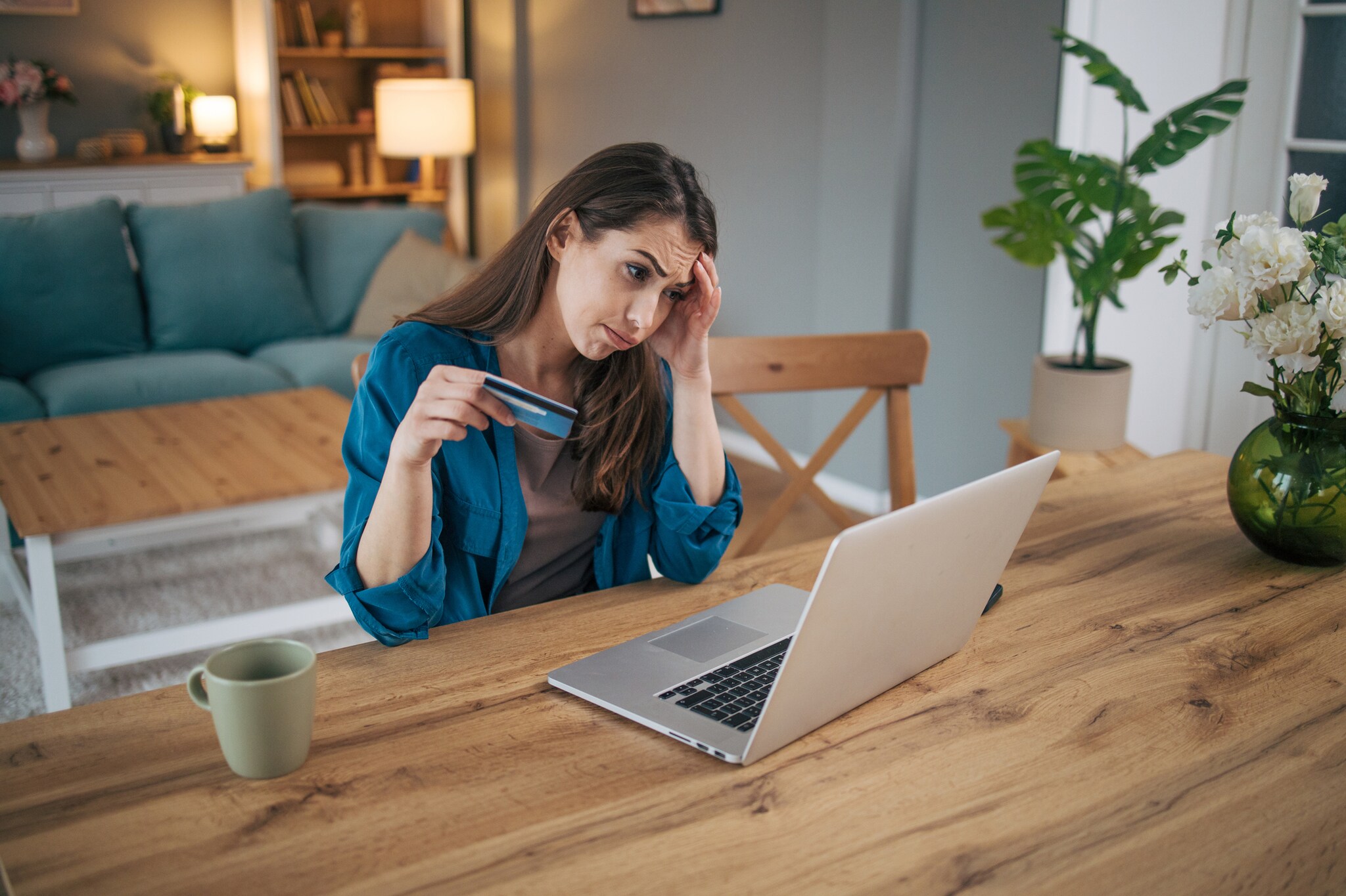 Eine Frau sitzt mit gerunzelter Stirn an einem Tisch vor einem Laptop. In der Hand hält sie eine blaue Karte.