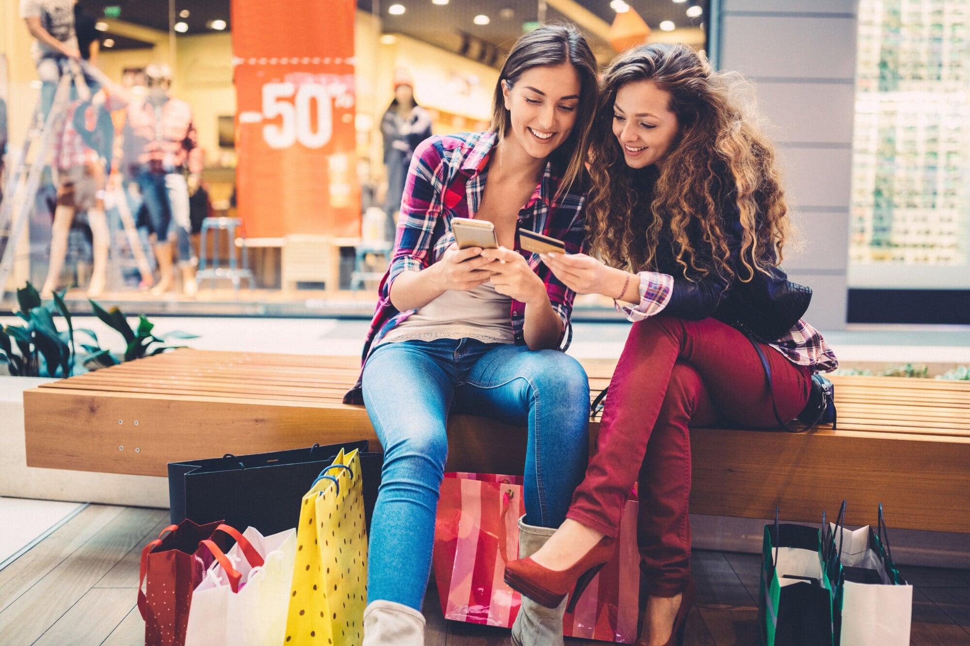 Zwei Personen sitzen in einer Shoppingmall auf einer Bank und schauen gemeinsam auf ein Smartphone und eine Kreditkarte. Zwei Personen sitzen in einer Shoppingmall auf einer Bank und schauen gemeinsam auf ein Smartphone und eine Kreditkarte.